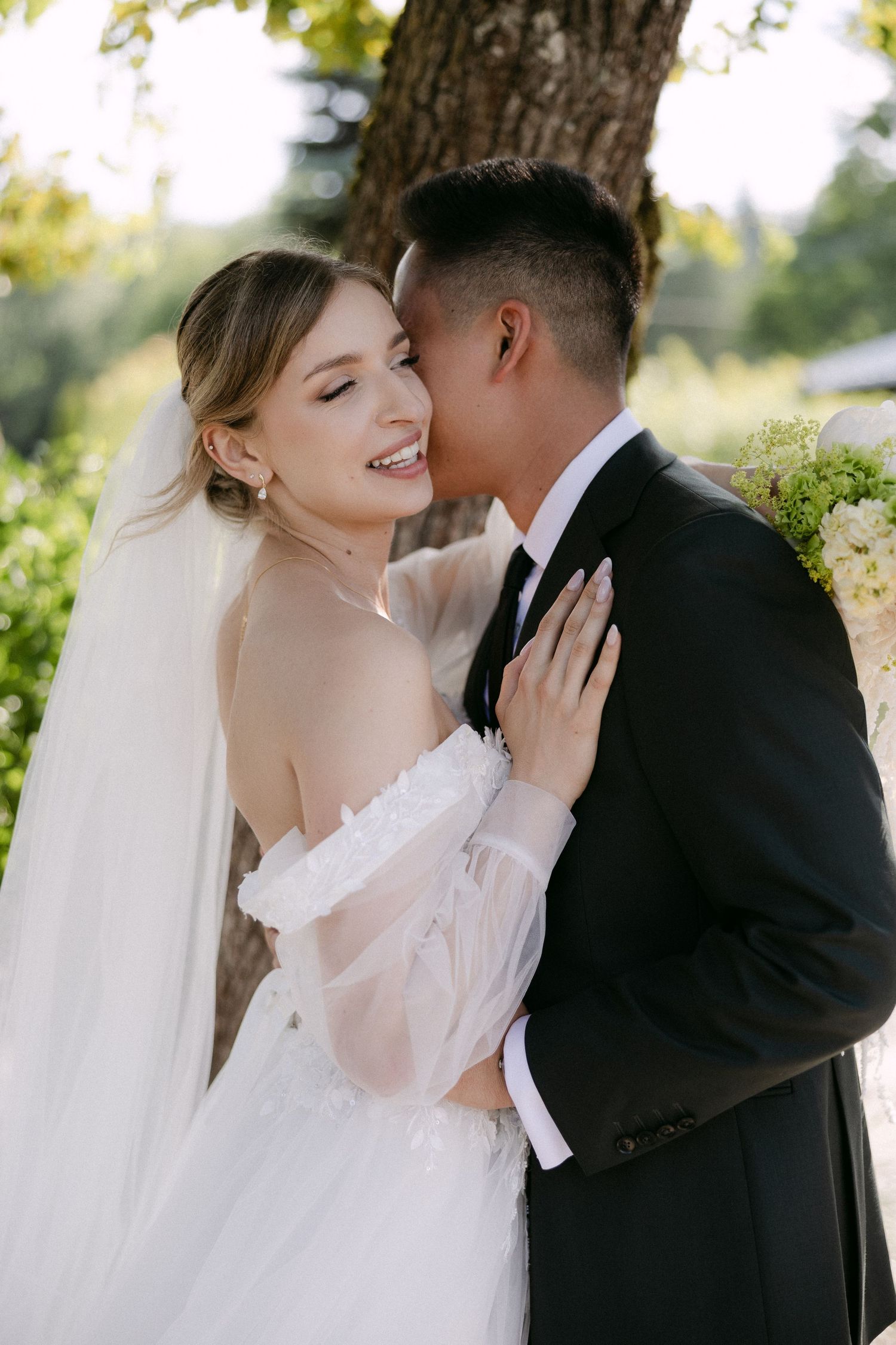 Groom kissing smiling bride on cheek outdoors in sunlit garden during romantic wedding portrait.