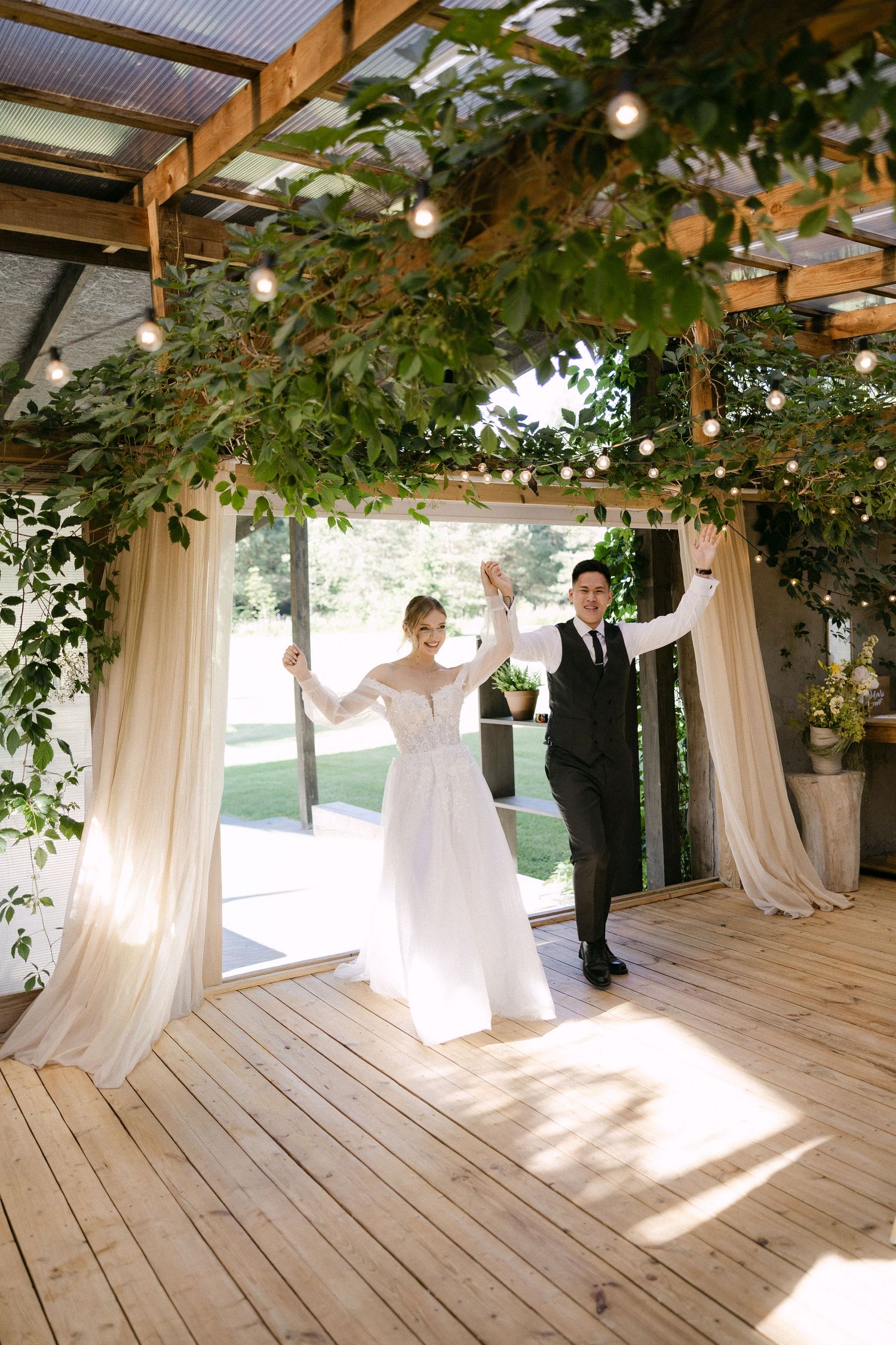 Newlyweds raise hands joyfully entering reception under greenery-draped wooden pergola.