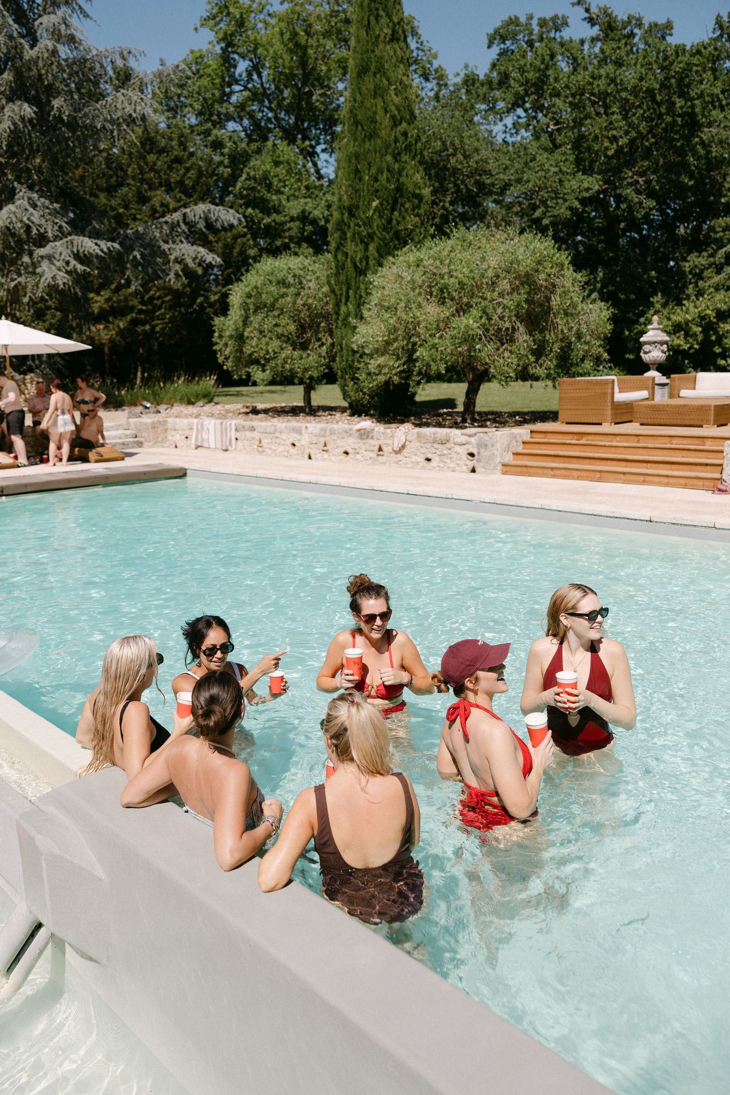 Women in swimsuits enjoying drinks in a sunny outdoor pool surrounded by lush green trees.