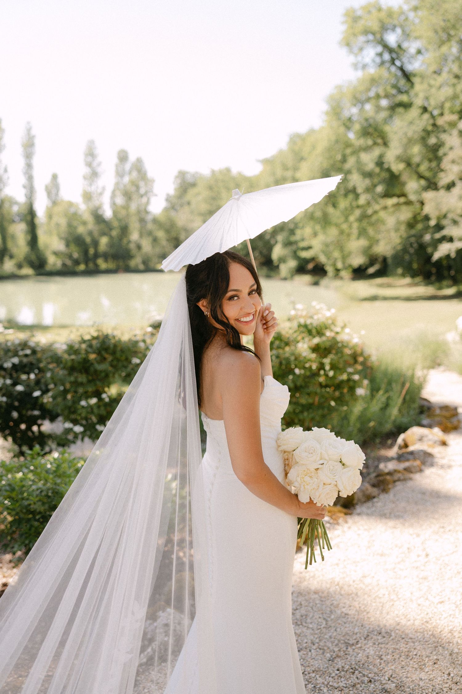 Smiling bride in strapless white gown holding bouquet and parasol beside a sunlit garden pond.