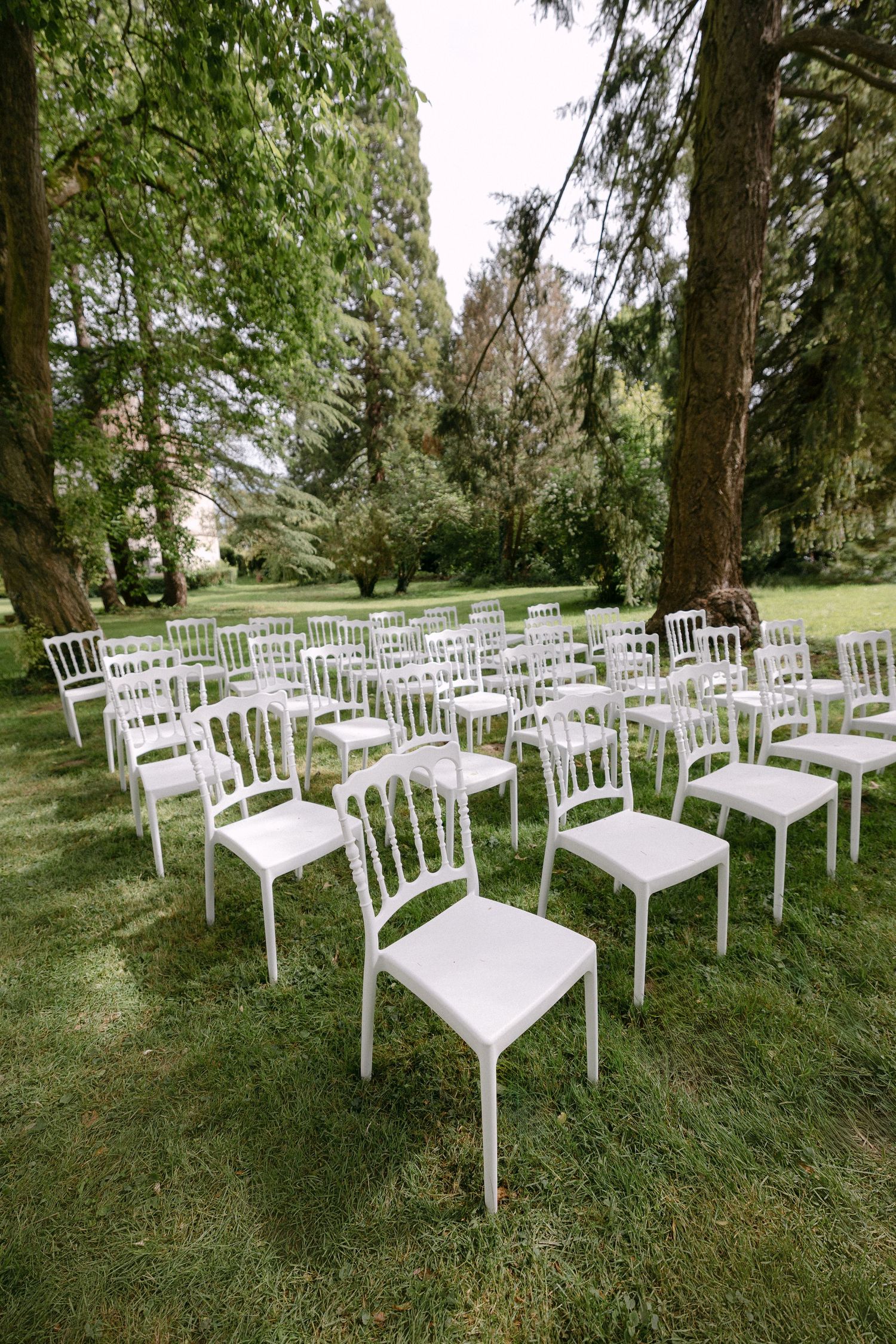 Rows of elegant white Napoleon chairs arranged on green grass for an outdoor garden wedding ceremony.