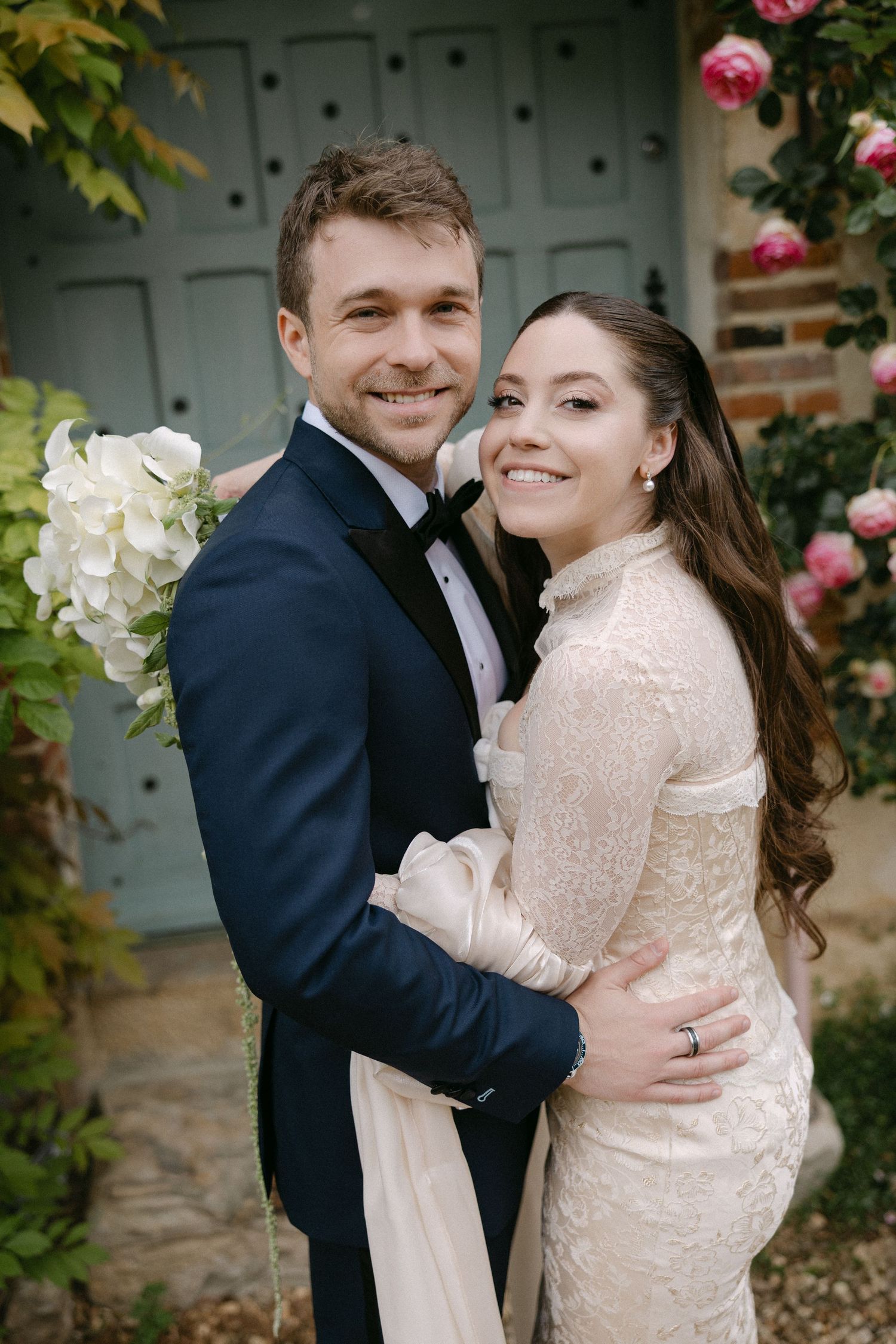 Happy bride and groom embracing outdoors, bride in lace long-sleeve gown, groom in navy tuxedo with bow tie.