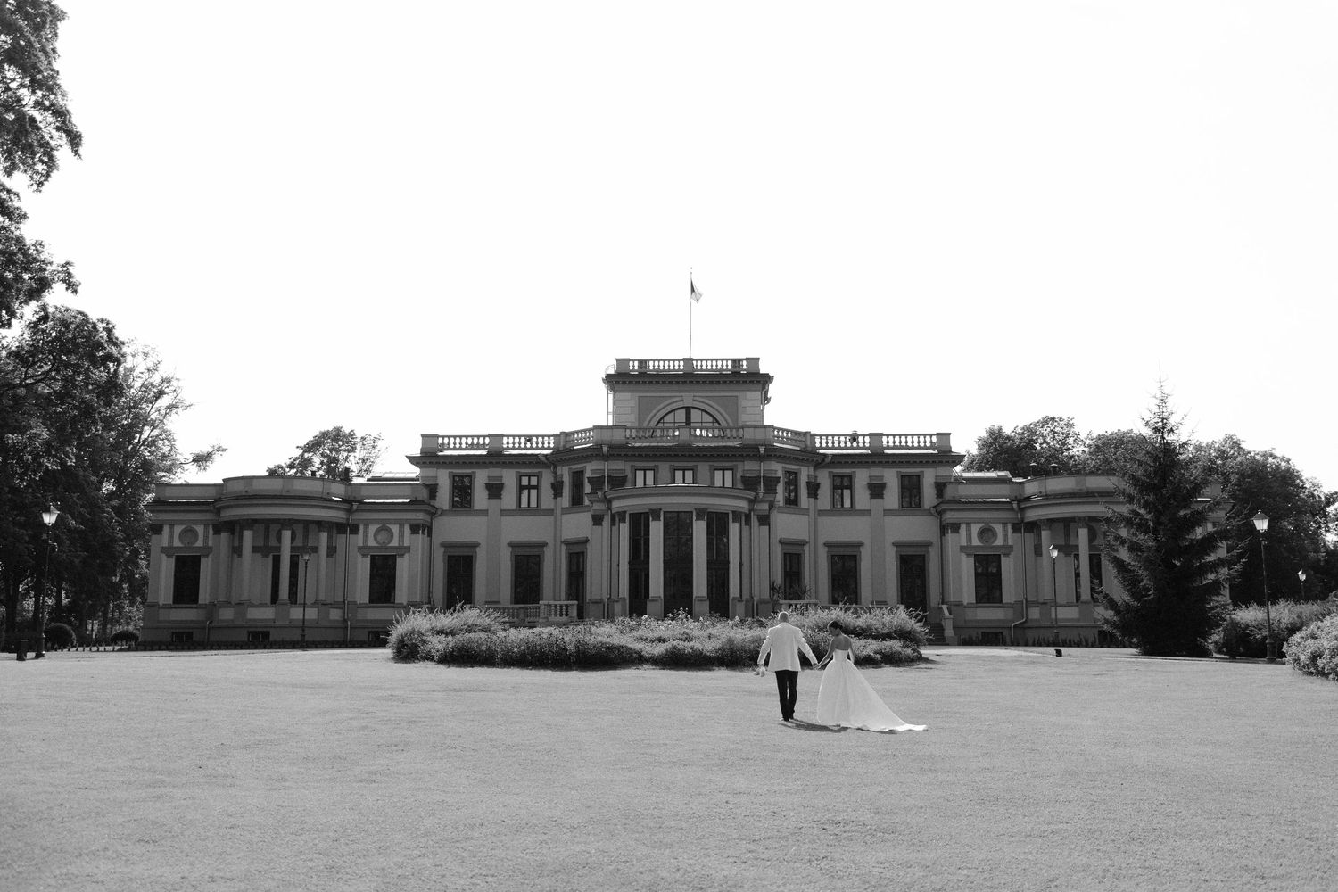 Black and white photo of a wedding couple walking toward a grand historic mansion on a lush green lawn.
