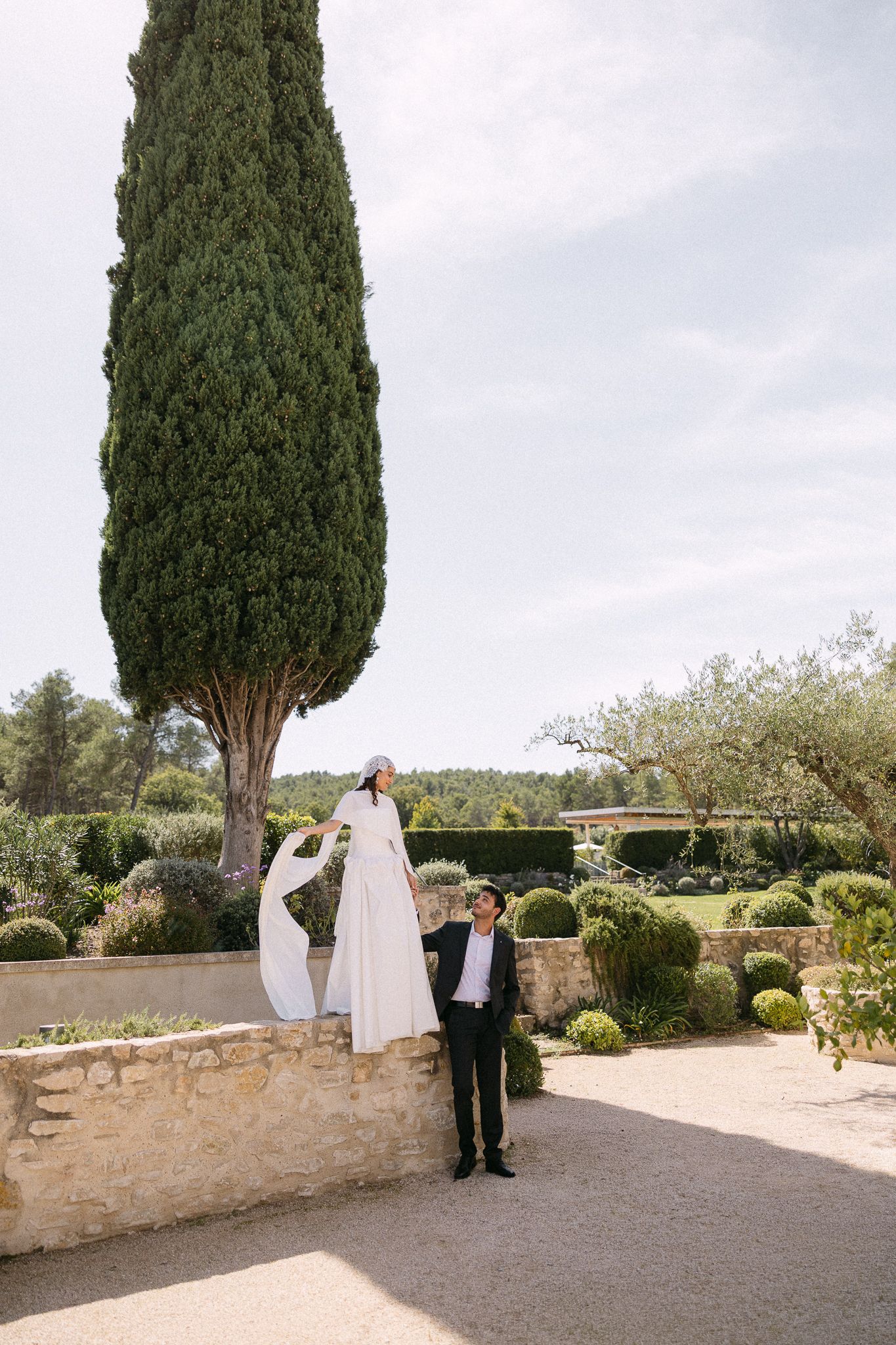 Bride standing on stone wall with groom below, white dress flowing, cypress tree backdrop in elegant garden setting.