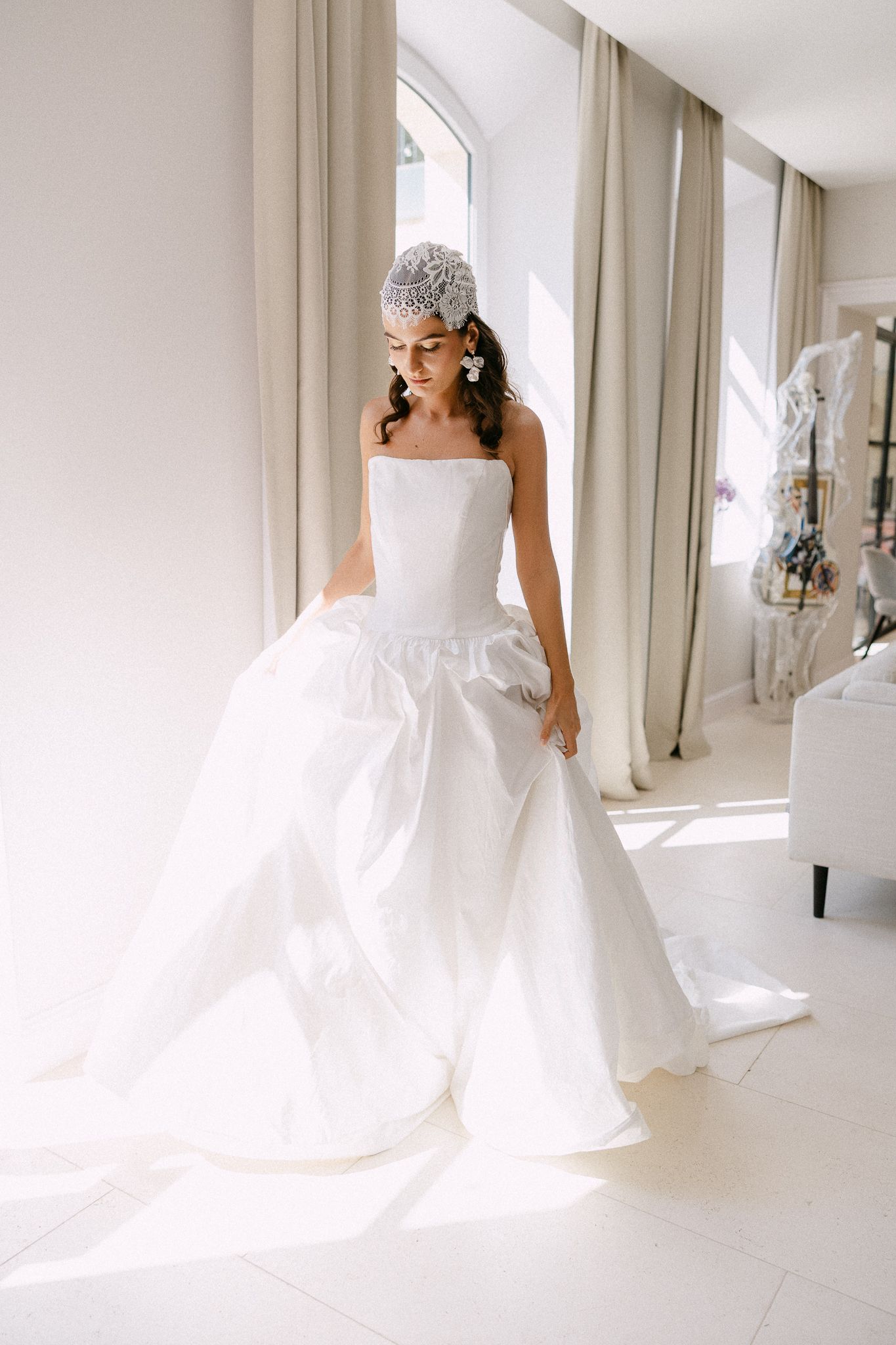 Bride in strapless white ballgown wedding dress with embellished headpiece standing in sunlit bridal suite.