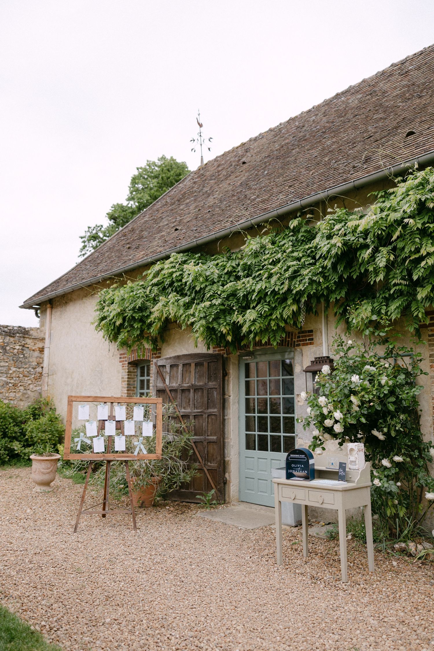 Rustic French stone cottage venue exterior with ivy, seating chart display, and welcome table on gravel path.