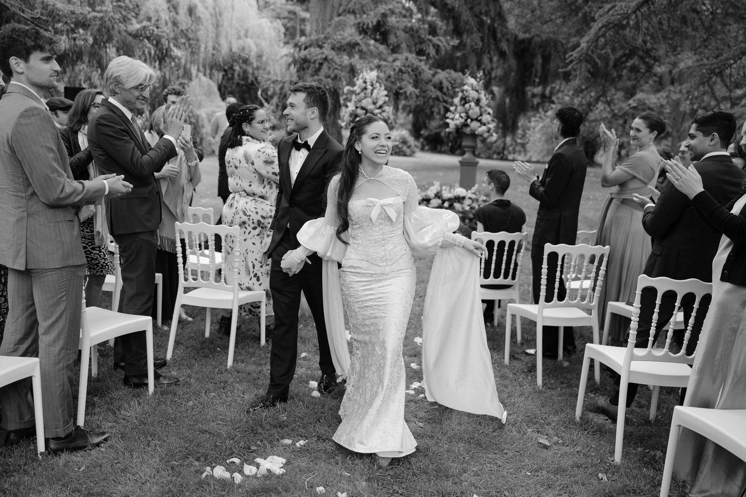 Newlyweds walk joyfully down outdoor aisle as guests applaud at garden wedding.