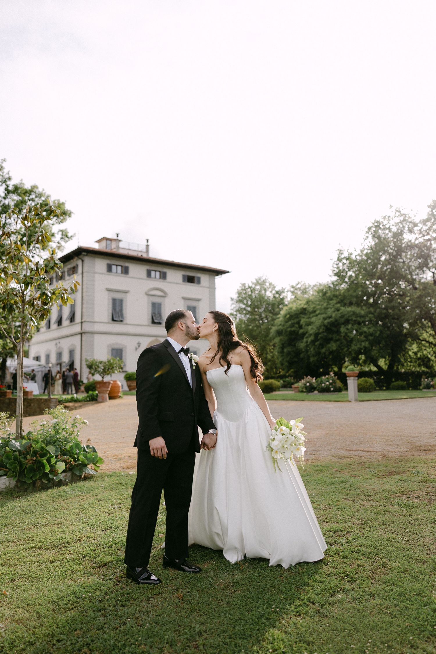 Bride and groom share a kiss on green lawn with grand Italian villa in background.