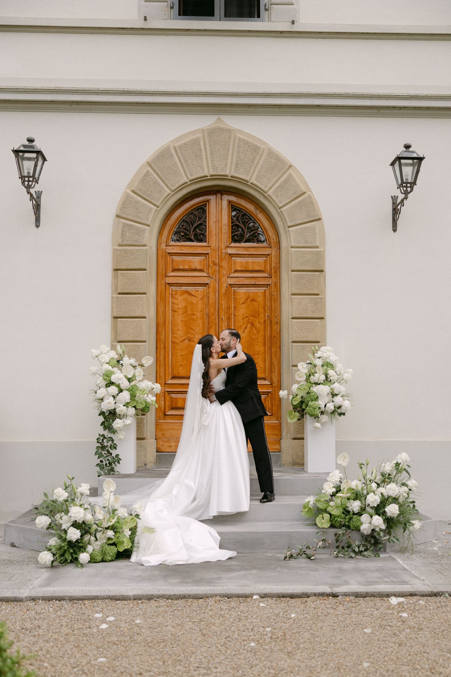 Bride and groom share first kiss on steps of elegant villa surrounded by white floral arrangements.