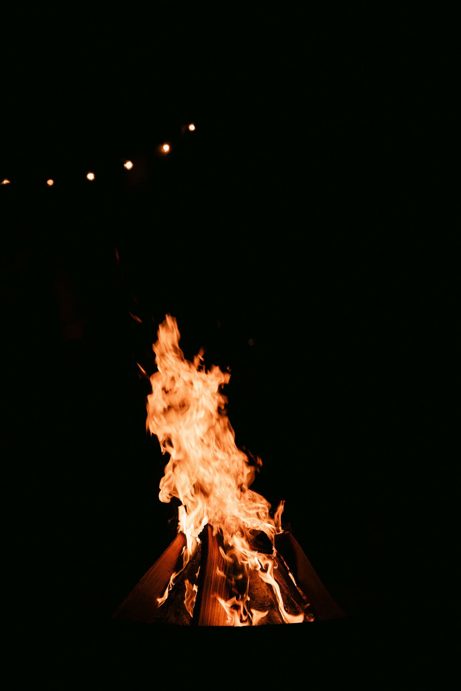 Dramatic bonfire burning brightly against a dark night sky with string lights visible in the background.