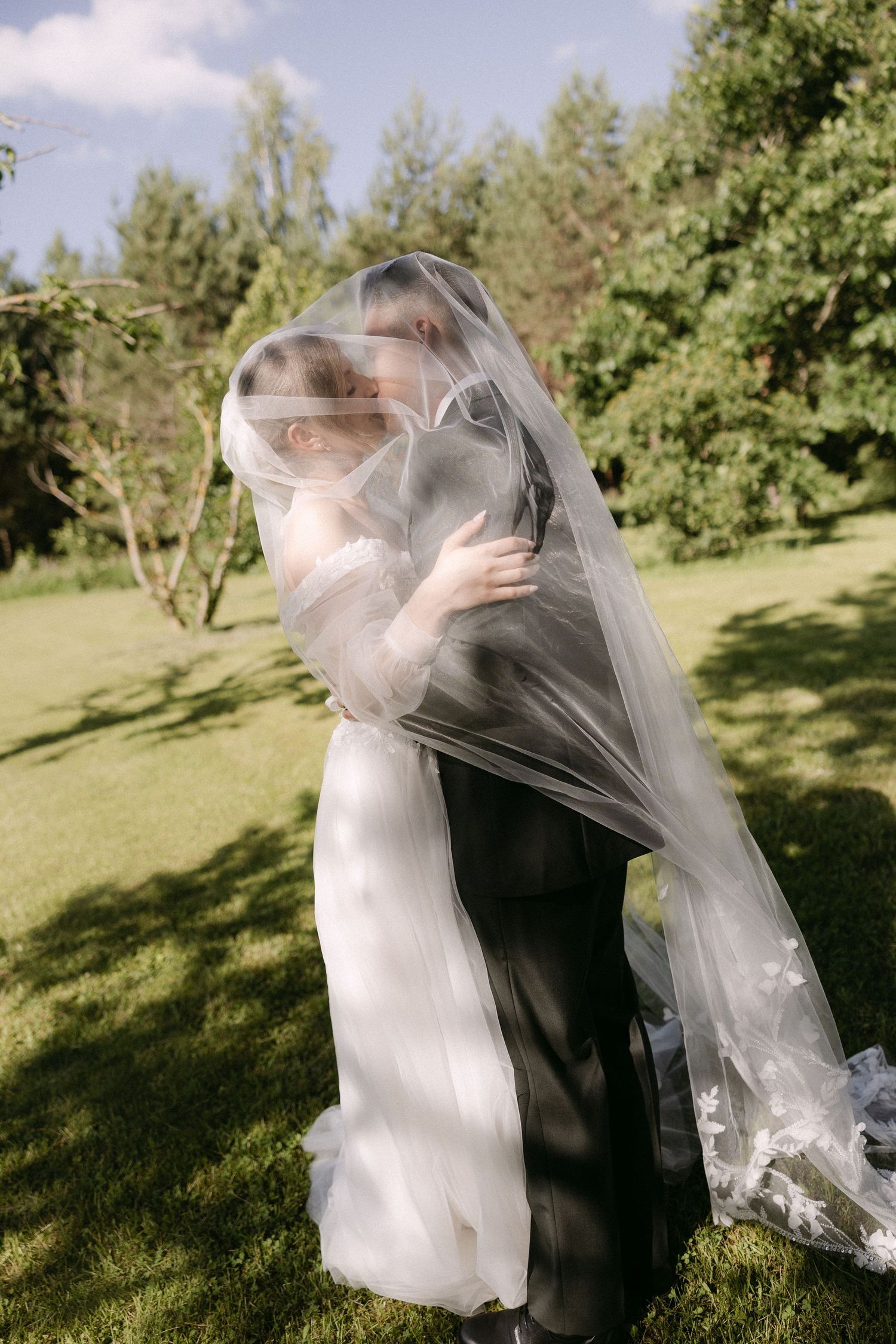 Bride and groom sharing a kiss hidden under a flowing wedding veil on a lush green lawn.