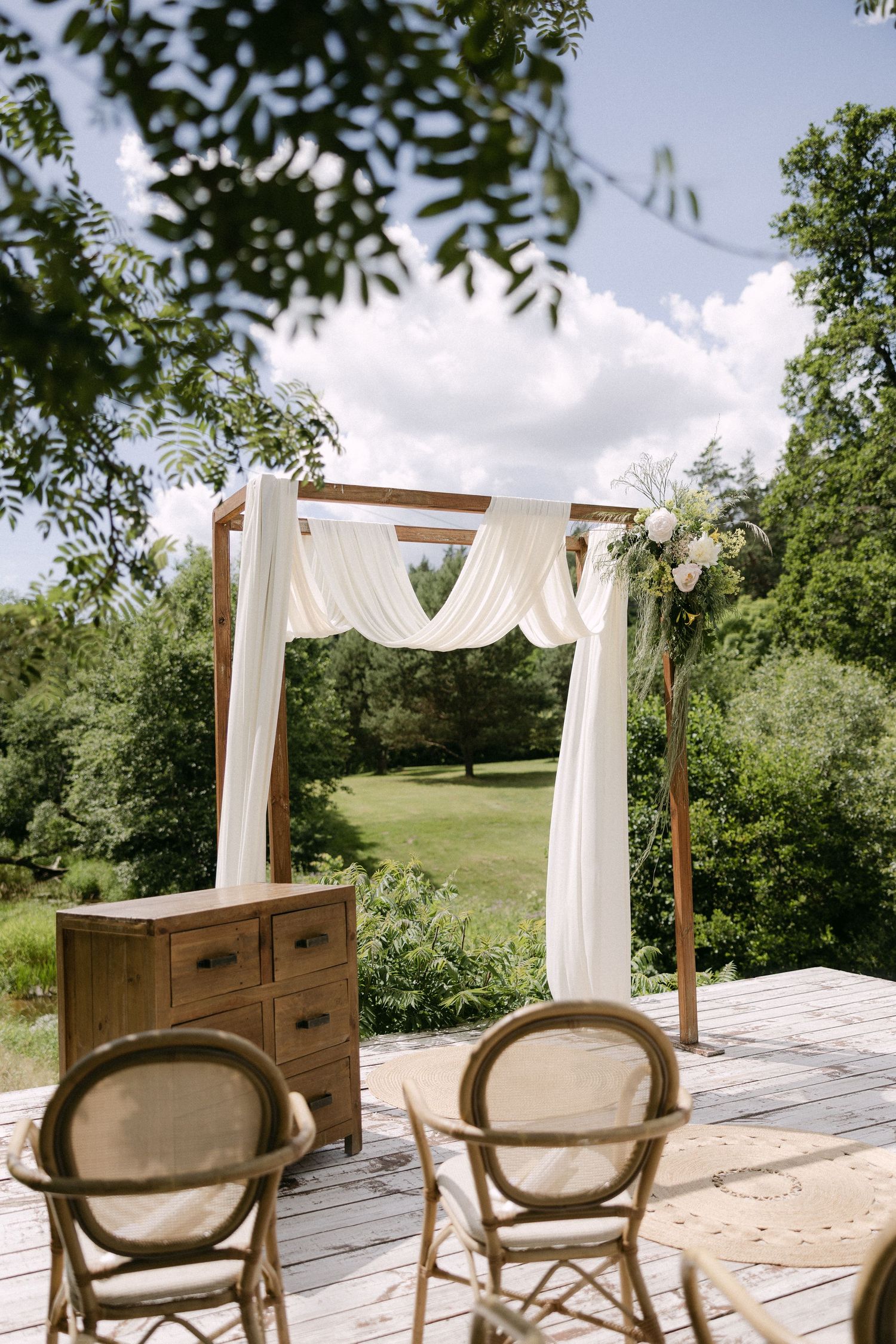 Outdoor wedding ceremony arch draped with white fabric and flowers on wooden deck surrounded by lush greenery.