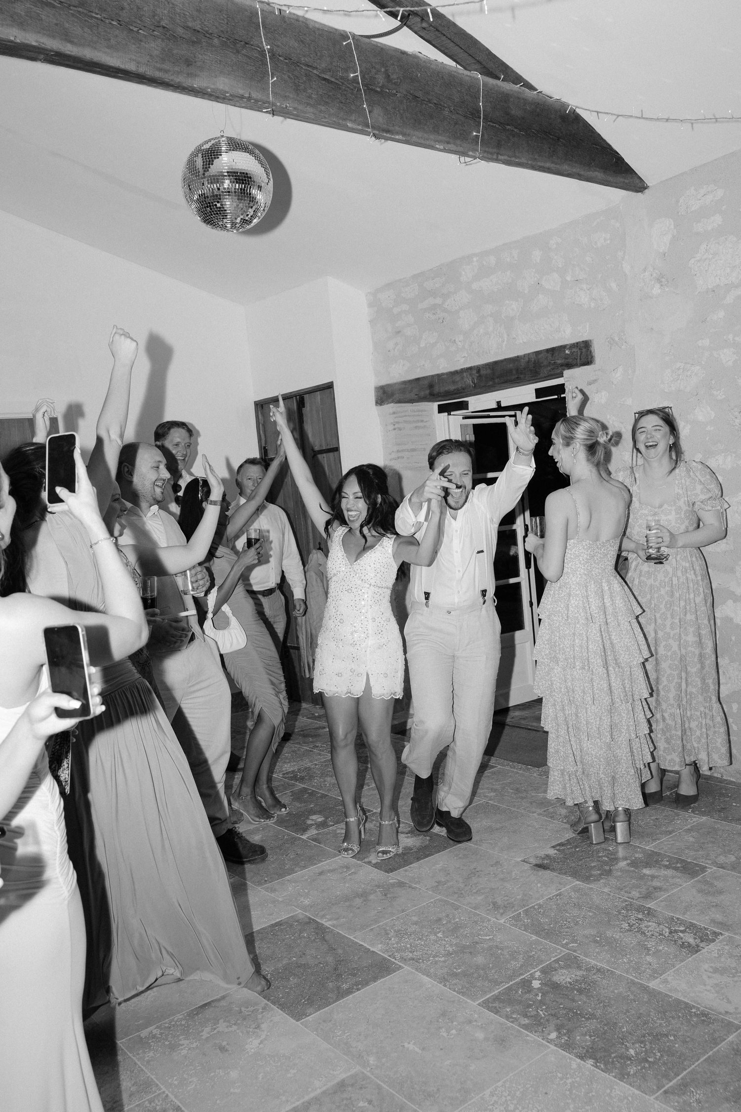 Black and white photo of bride and groom dancing joyfully with guests at wedding reception.