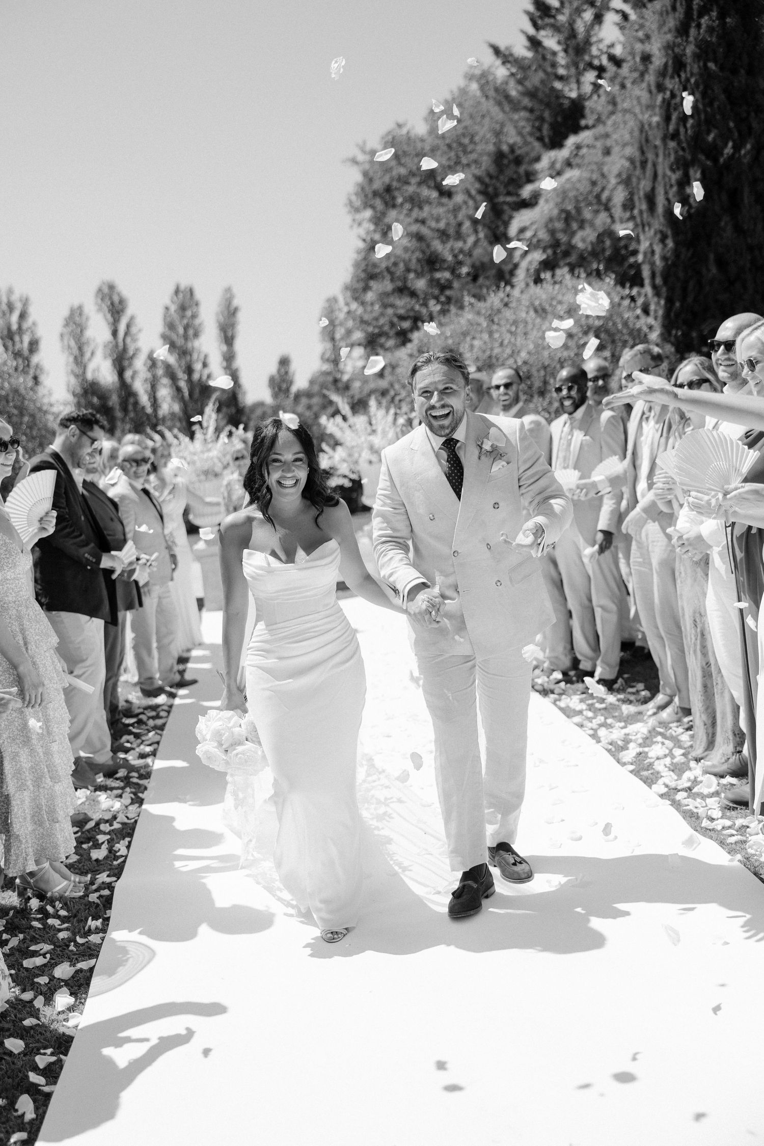 Joyful newlyweds walk down the aisle as guests toss flower petals in a sunny outdoor wedding ceremony.