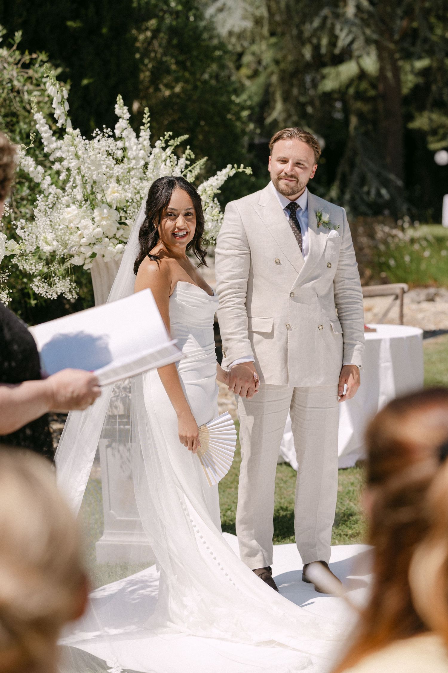 Joyful bride and groom holding hands at outdoor wedding ceremony with white floral backdrop.