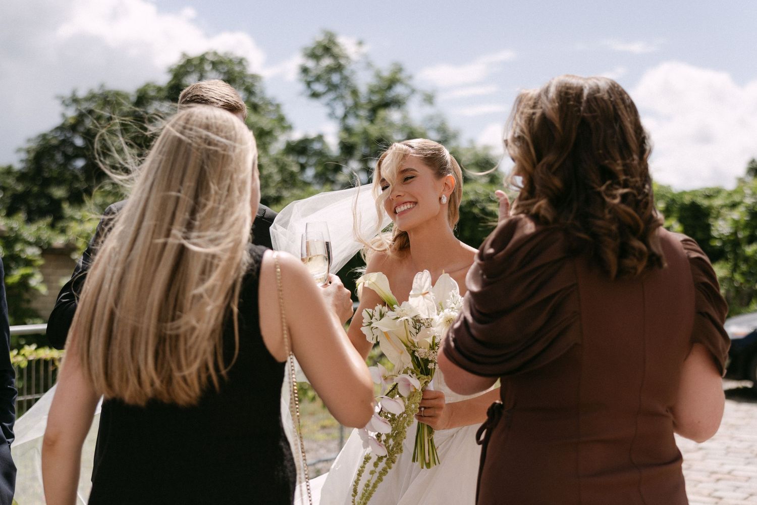 Smiling bride in a white gown holds a bouquet while greeting guests outdoors on her wedding day.