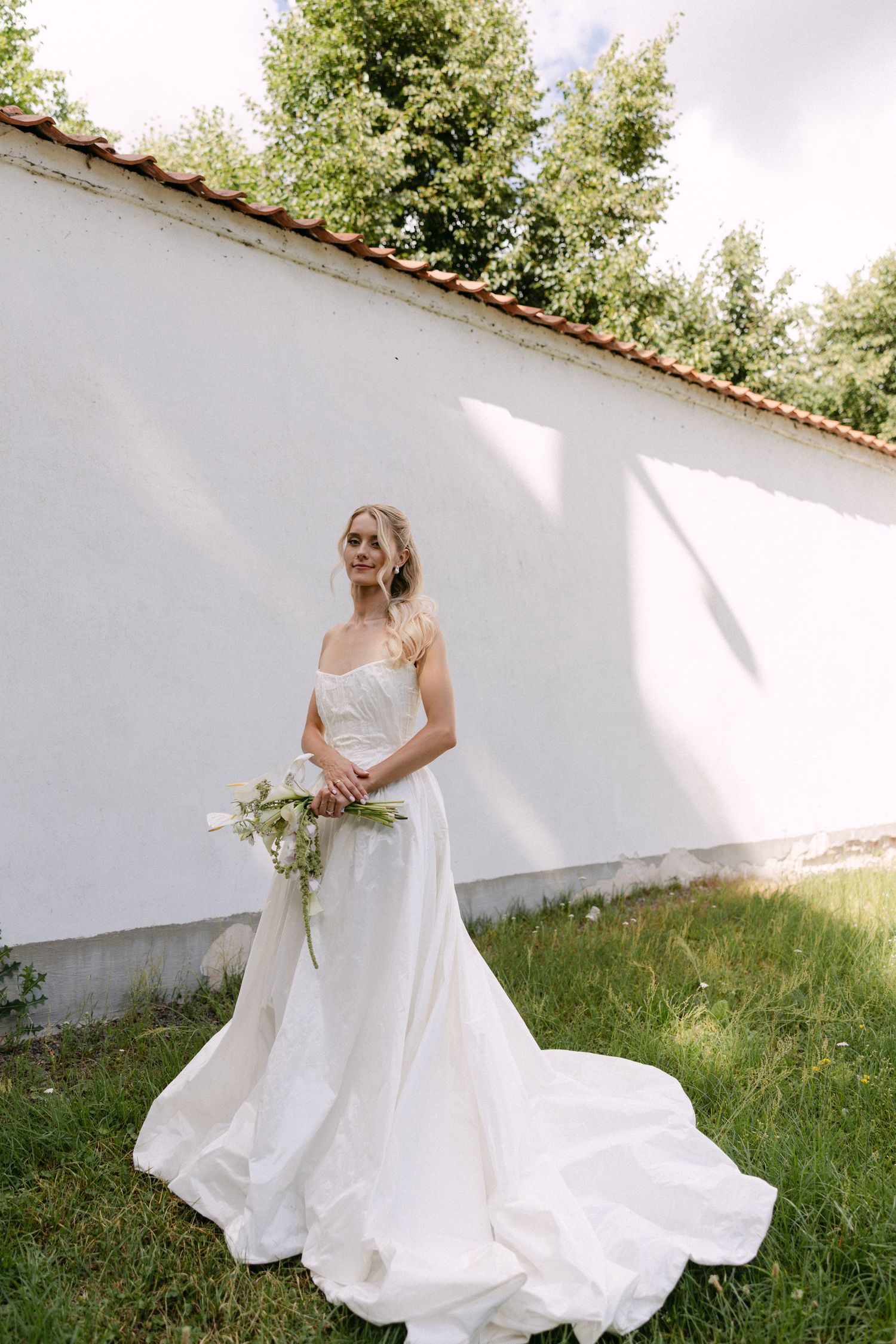 Blonde bride in a flowing white A-line gown holds a bouquet outdoors near a white wall with lush green trees.
