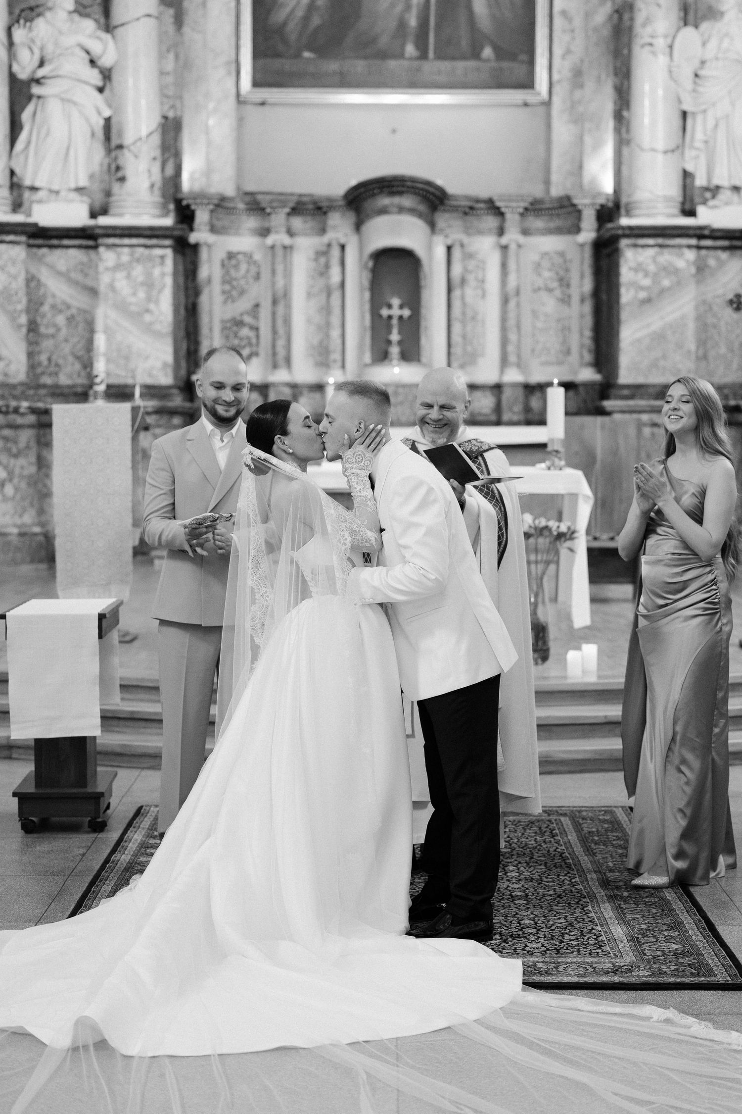 Bride and groom sharing first kiss at church altar surrounded by wedding party in black and white.