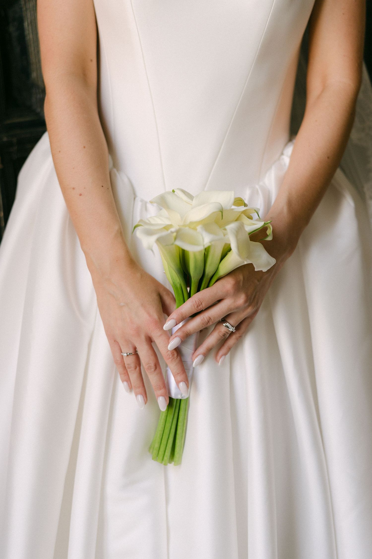 Close-up of bride holding white calla lily bouquet against satin wedding gown with manicured hands.