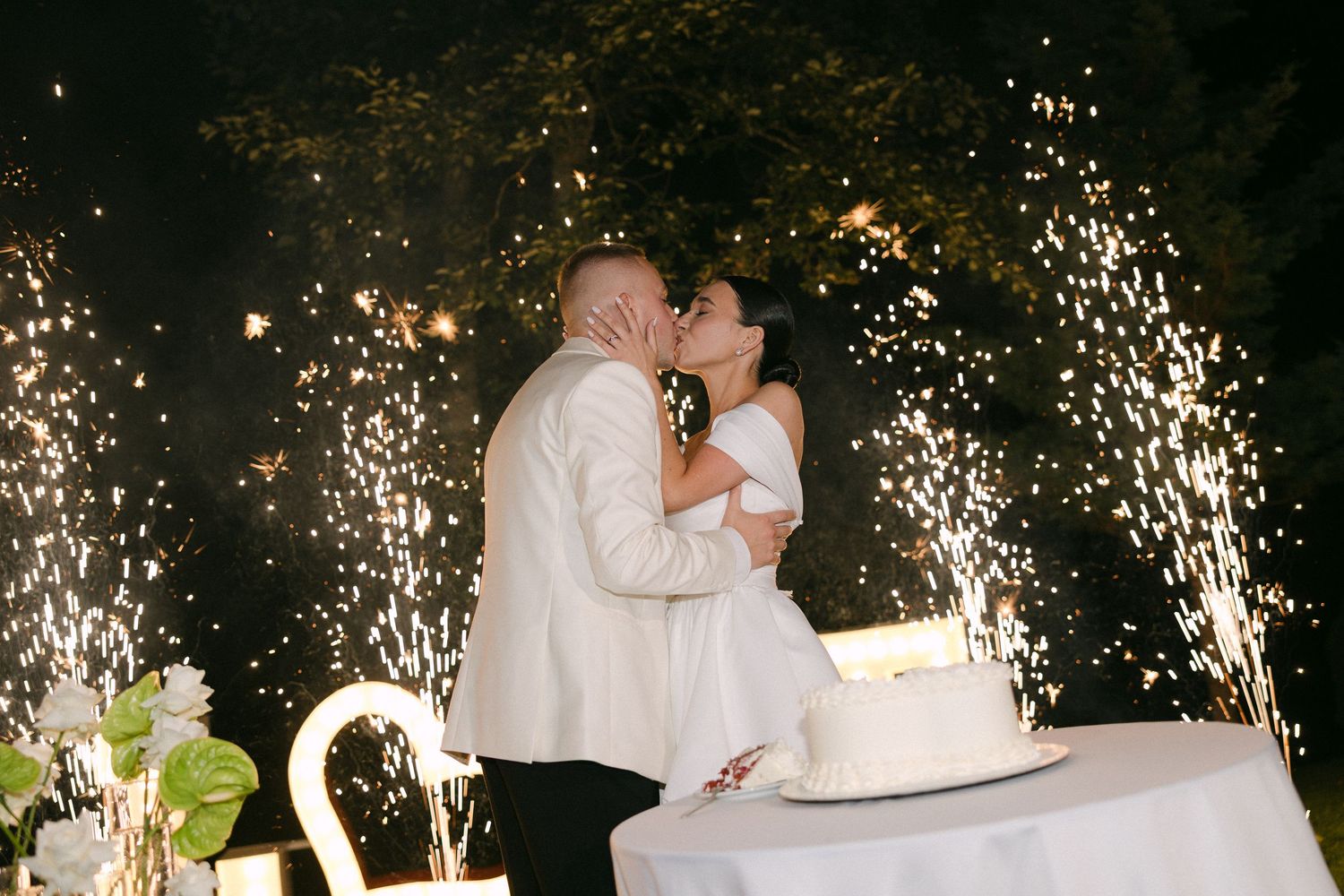 Bride and groom sharing a romantic kiss while cutting their wedding cake surrounded by sparkling fireworks.