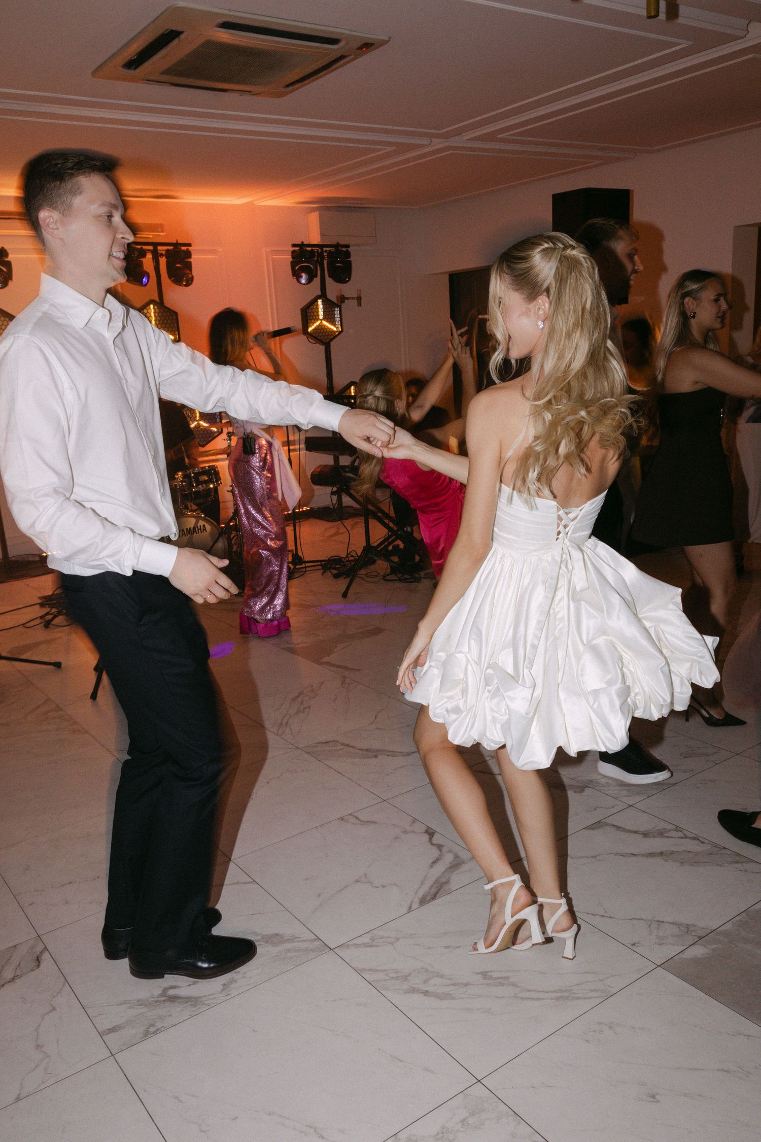 Bride in short white ruffled dress dancing energetically with groom at wedding reception.