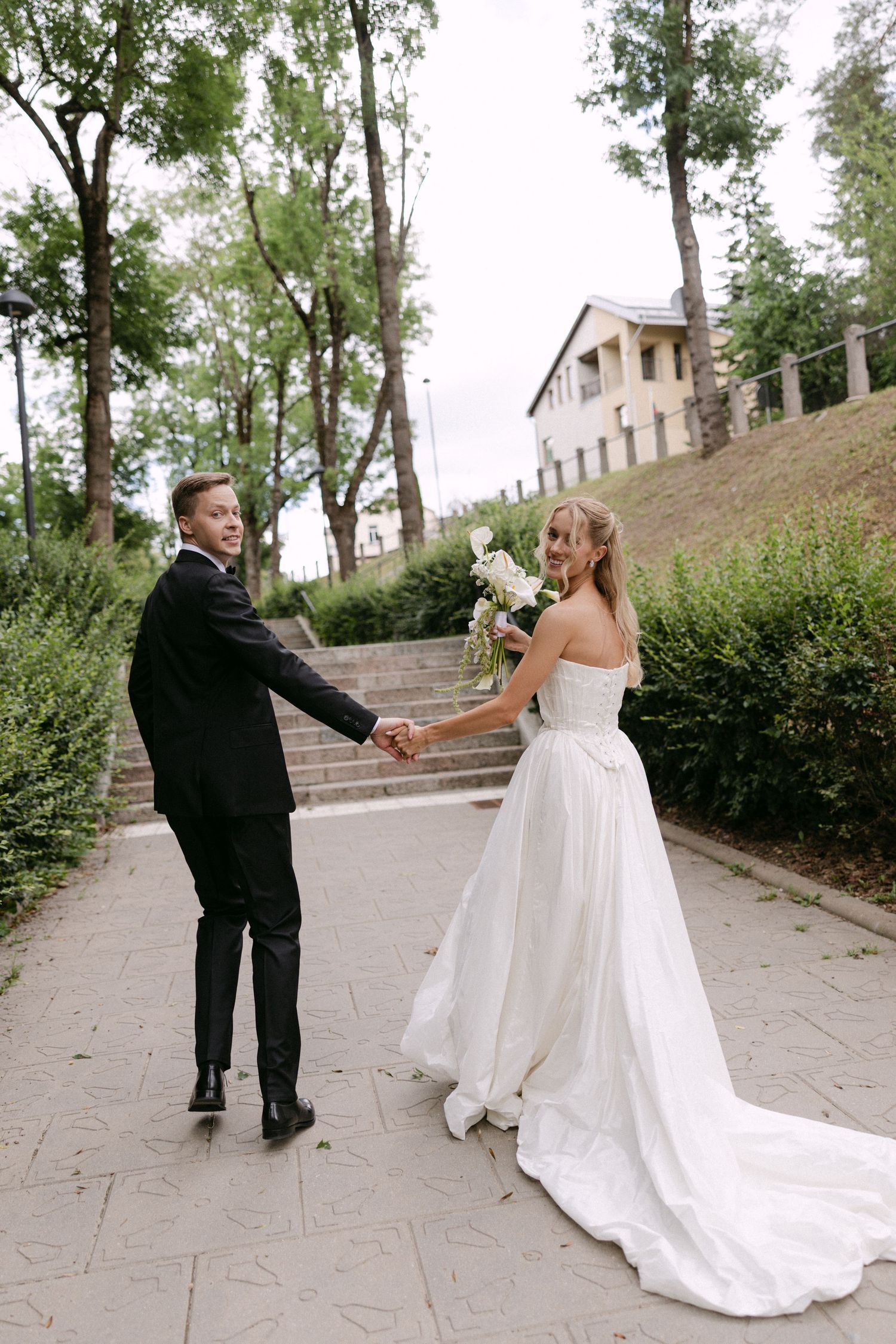 Newlyweds walk hand in hand on a tree-lined path, bride in white gown with long train.