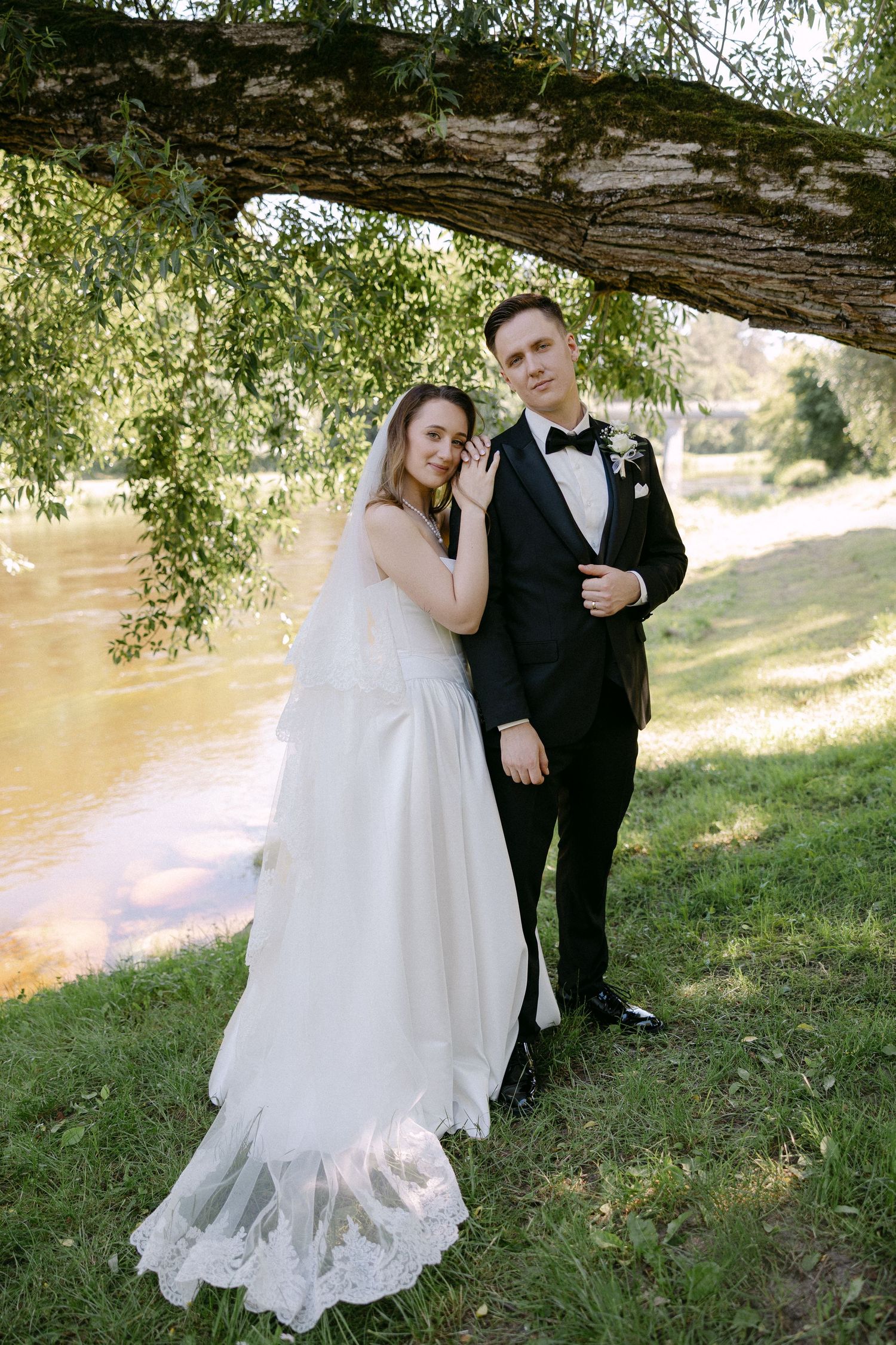 Bride in white lace gown and groom in black tuxedo posing under a tree by a river on their wedding day.