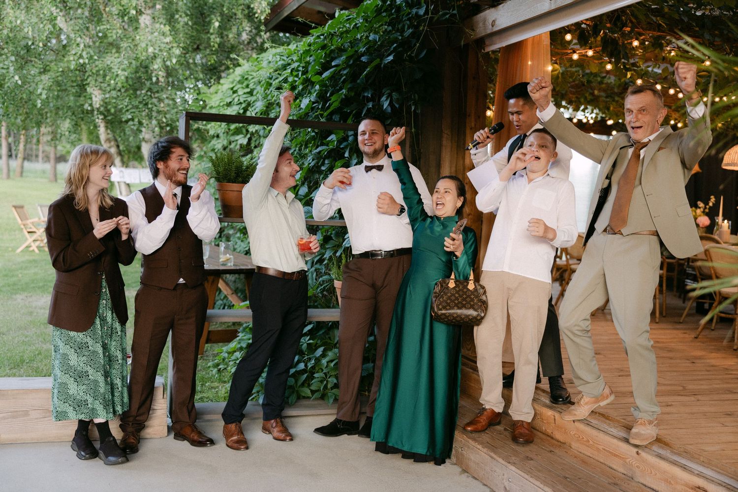 Wedding party group celebrates and cheers outside rustic venue with string lights overhead.