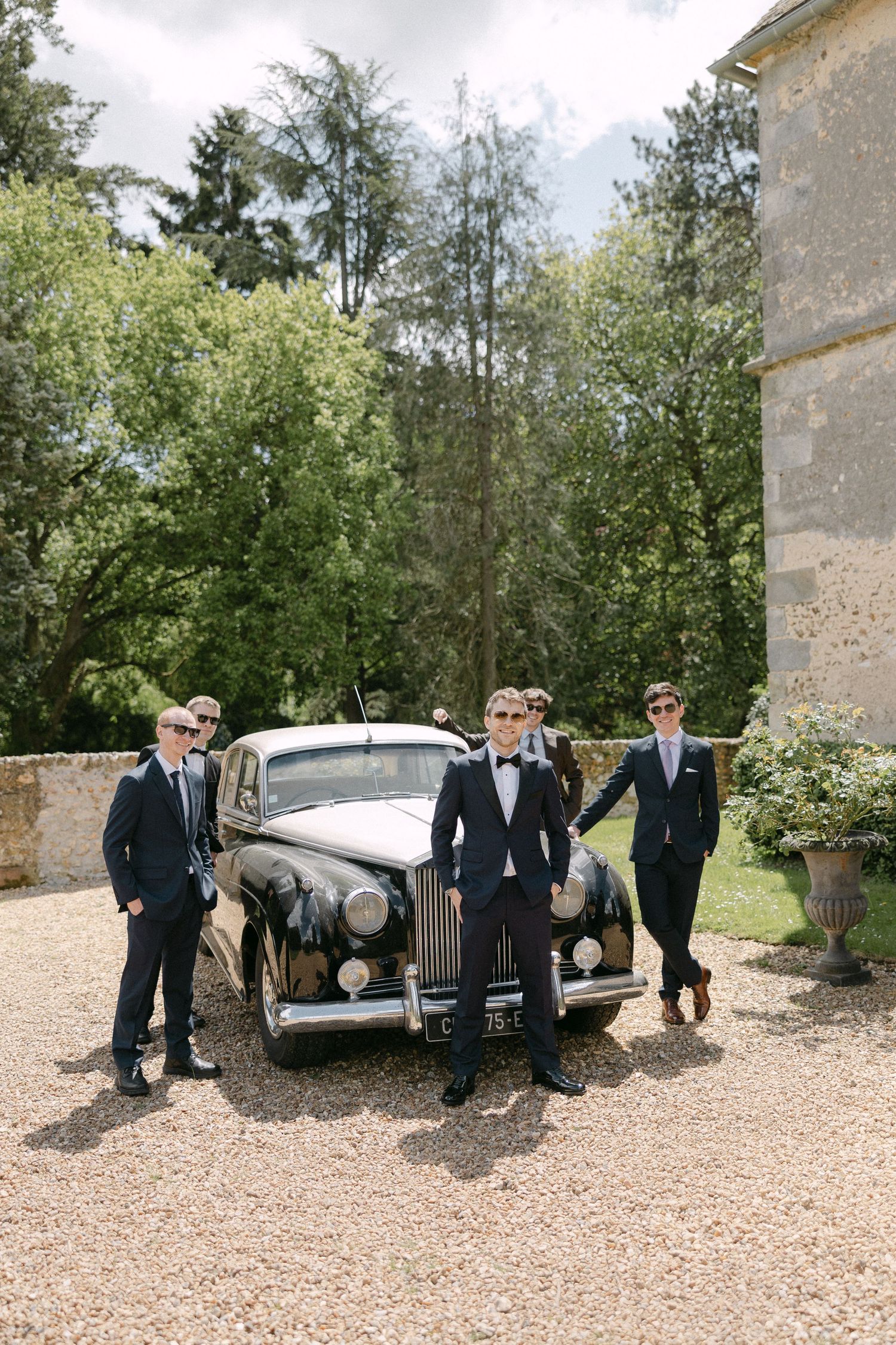 Four groomsmen in black suits and sunglasses posing with a vintage classic car on a gravel driveway.