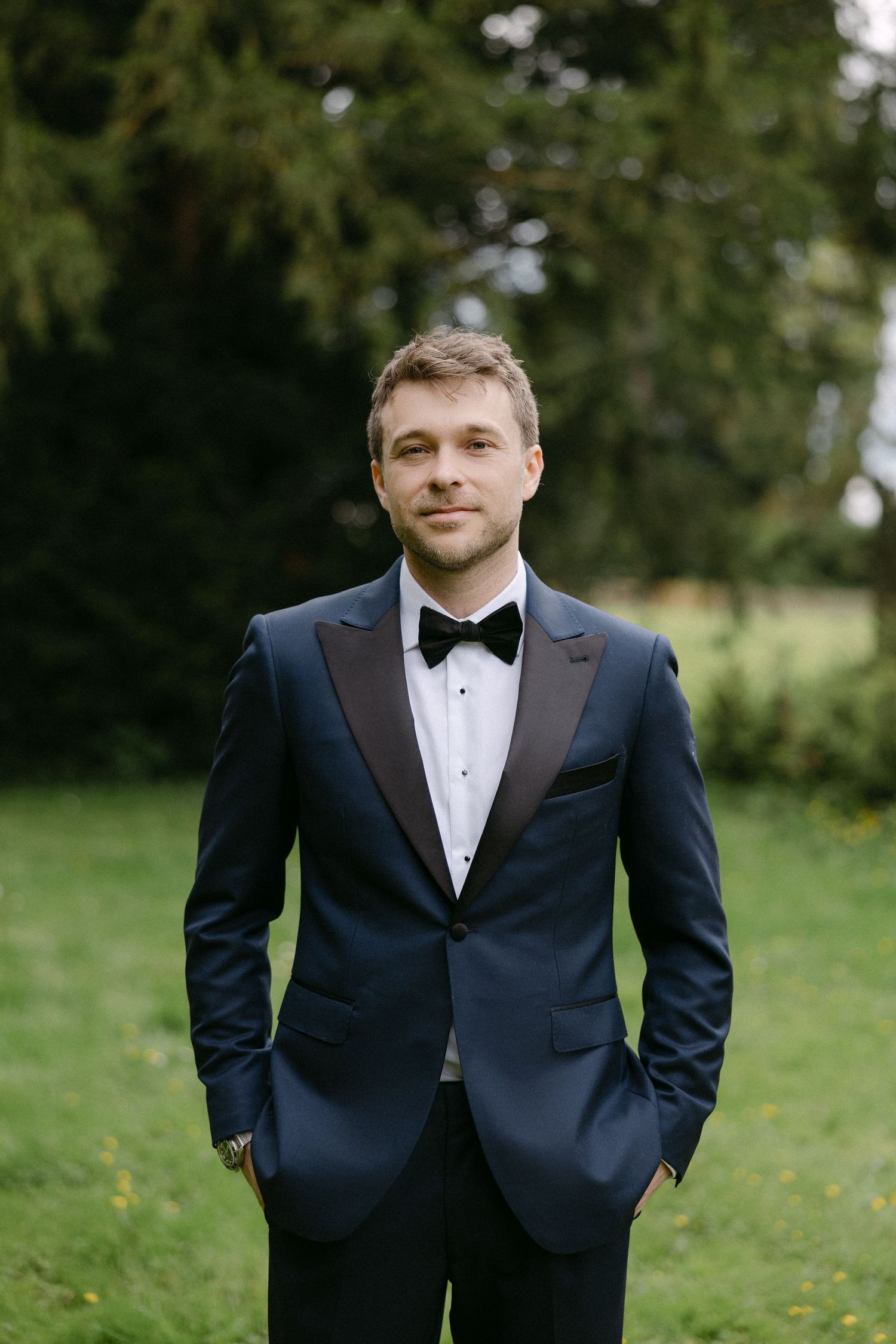 Groom in navy tuxedo with black bow tie standing on green lawn at outdoor wedding celebration.