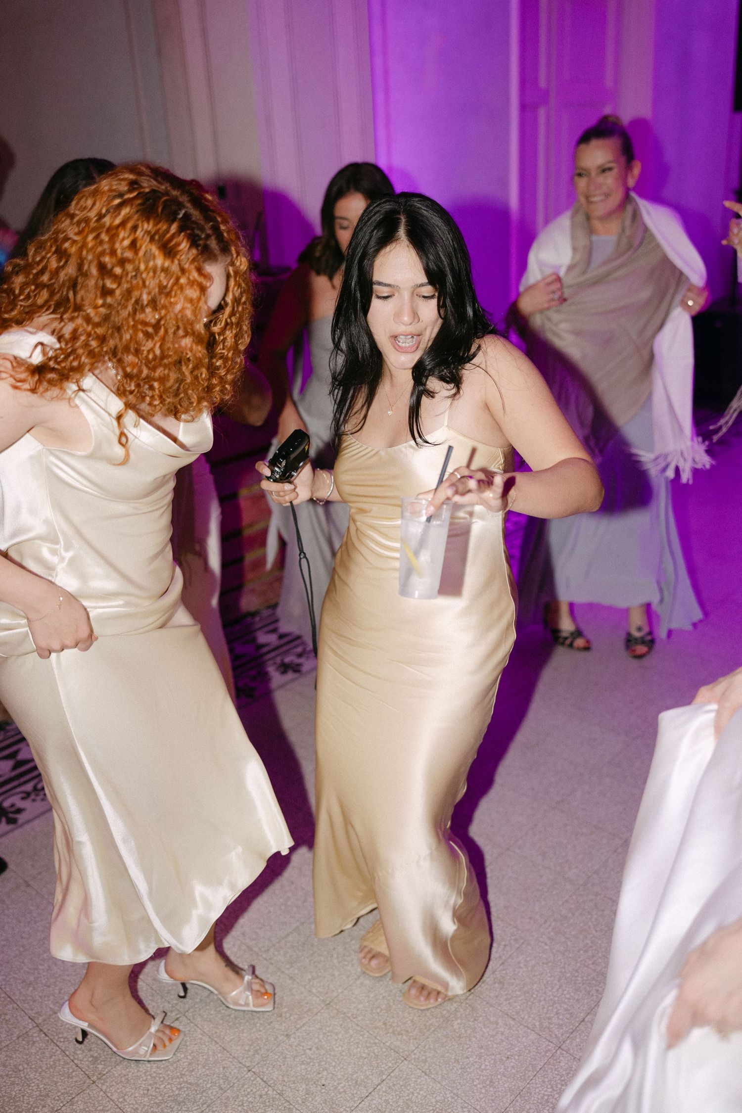 Bridesmaids in gold satin dresses dance energetically on wedding reception dance floor under purple lights.