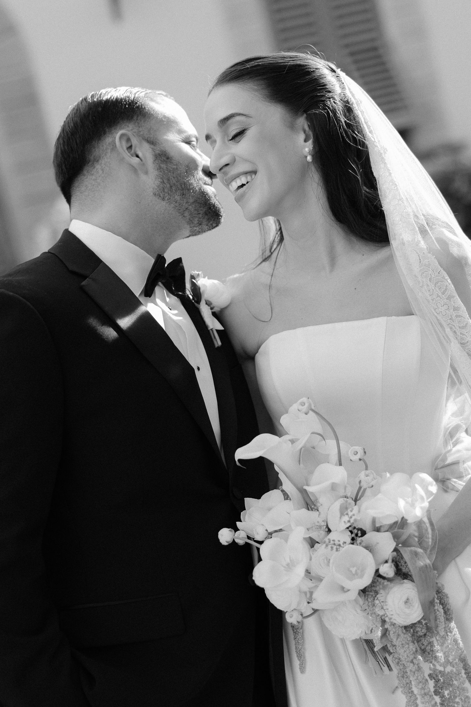 Black and white photo of smiling bride and groom sharing an intimate moment, bride holding white floral bouquet.