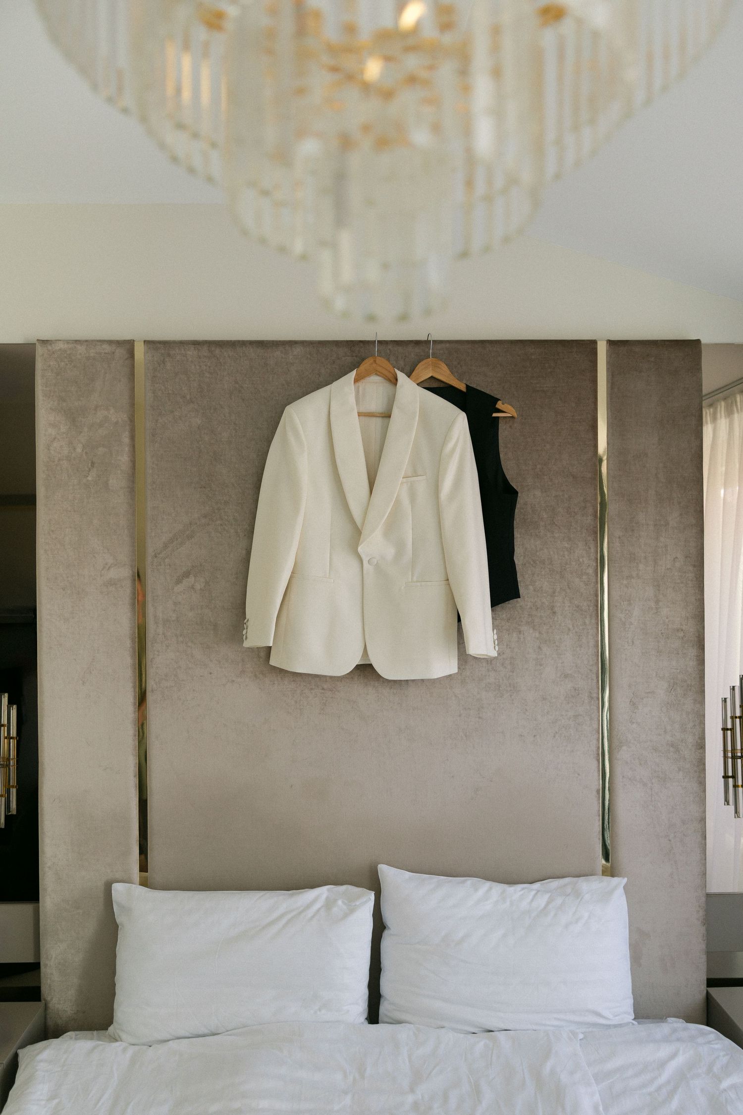 White tuxedo jacket and black vest hanging on bed headboard beneath elegant crystal chandelier in luxury hotel room.