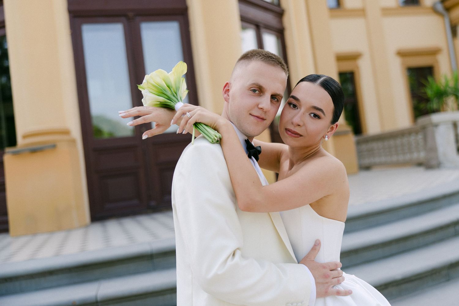 Bride and groom embracing outdoors in front of a yellow building holding a green bouquet.