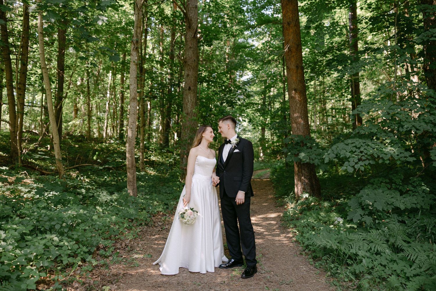 Bride and groom share romantic moment on forest path in wedding attire.