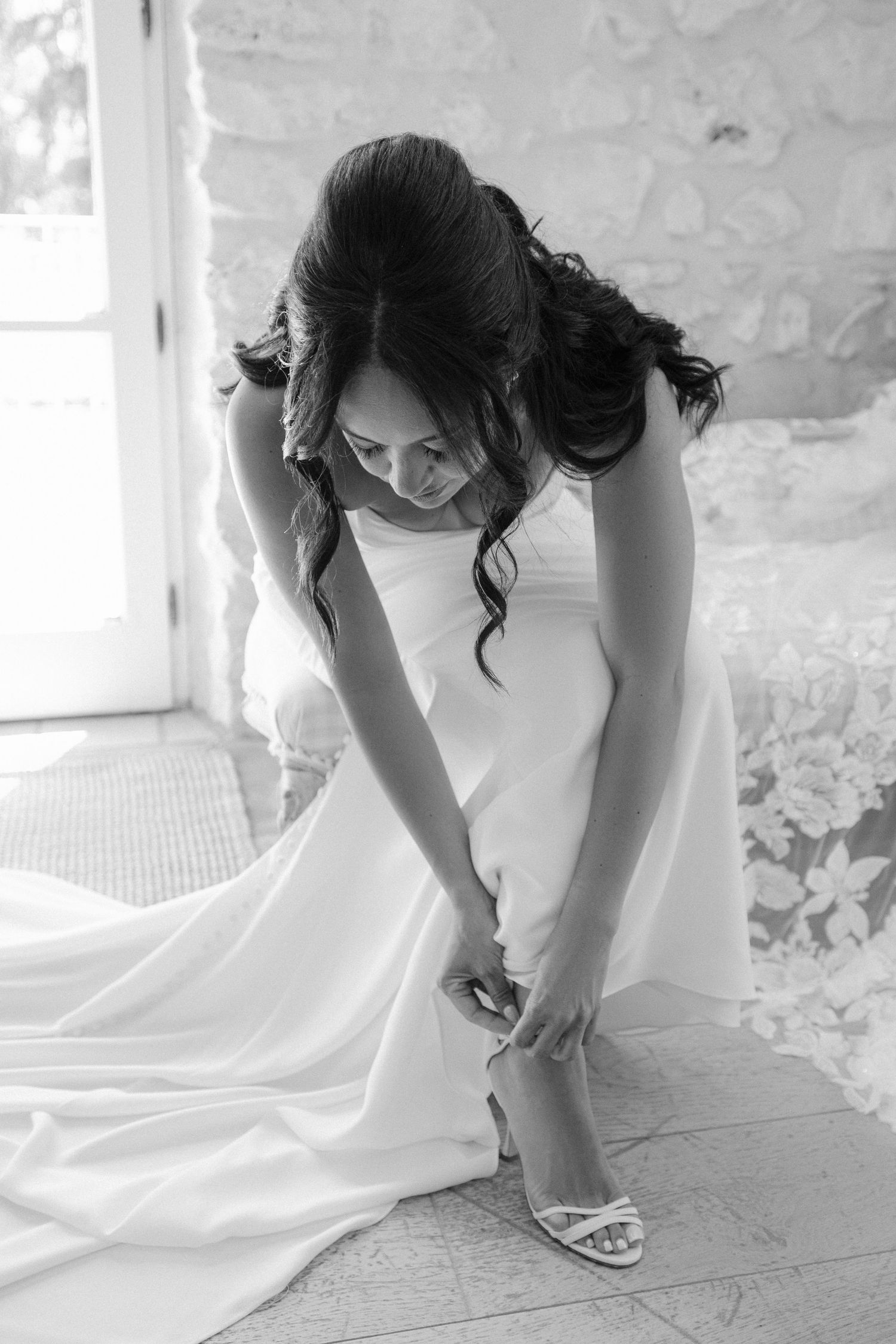 Bride in white gown fastening strappy heels in black and white bridal preparation photo.