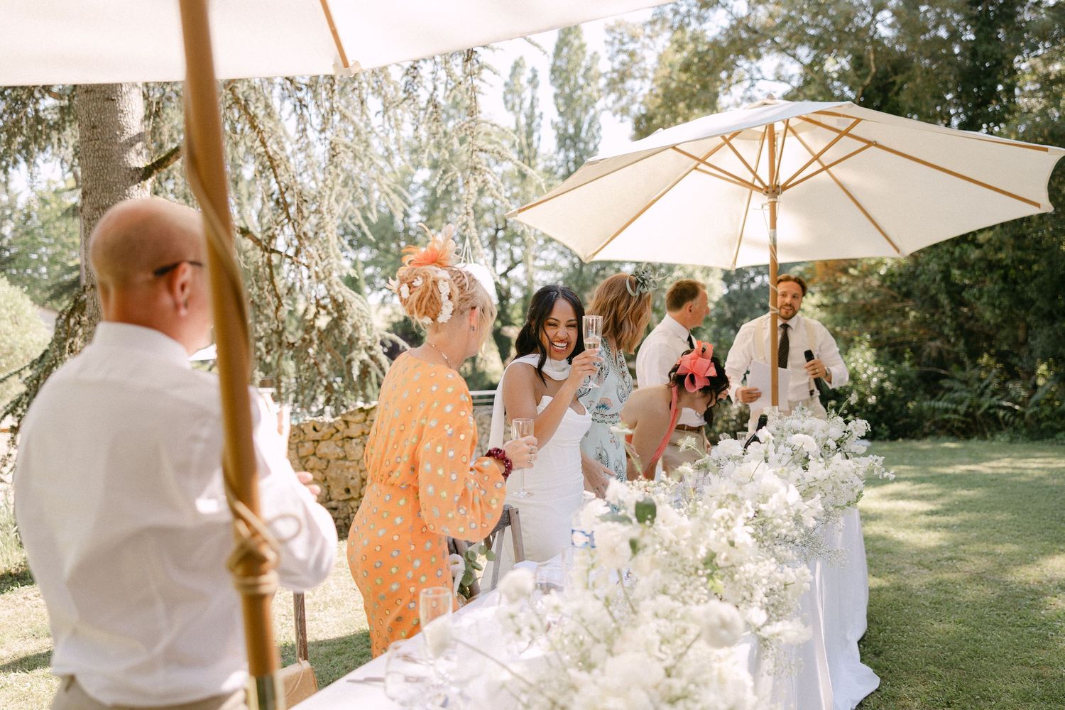 Elegant outdoor wedding cocktail hour with white floral table, umbrellas, and guests mingling.