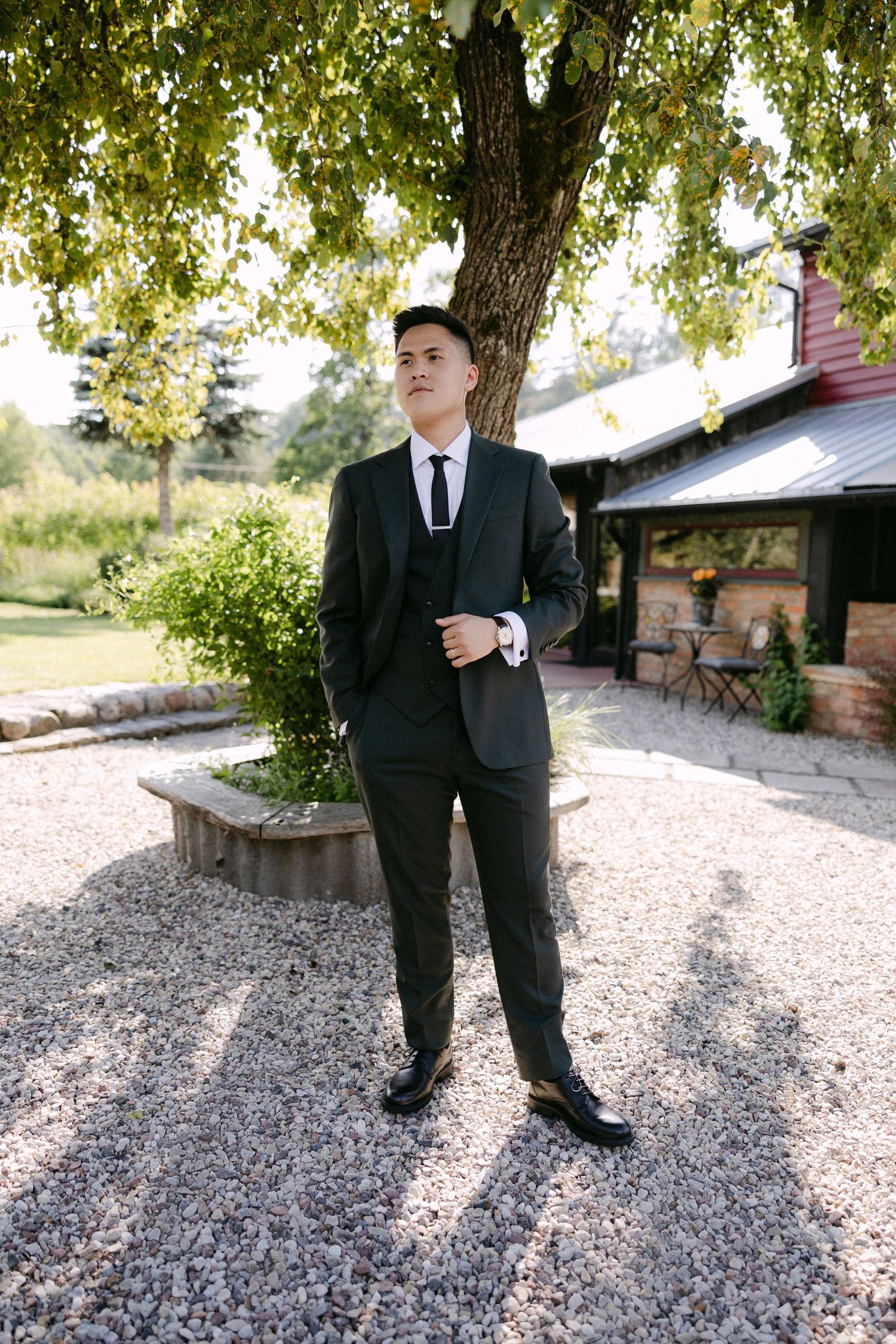 Groom in dark suit stands confidently on gravel path beneath a tree outside rustic venue.