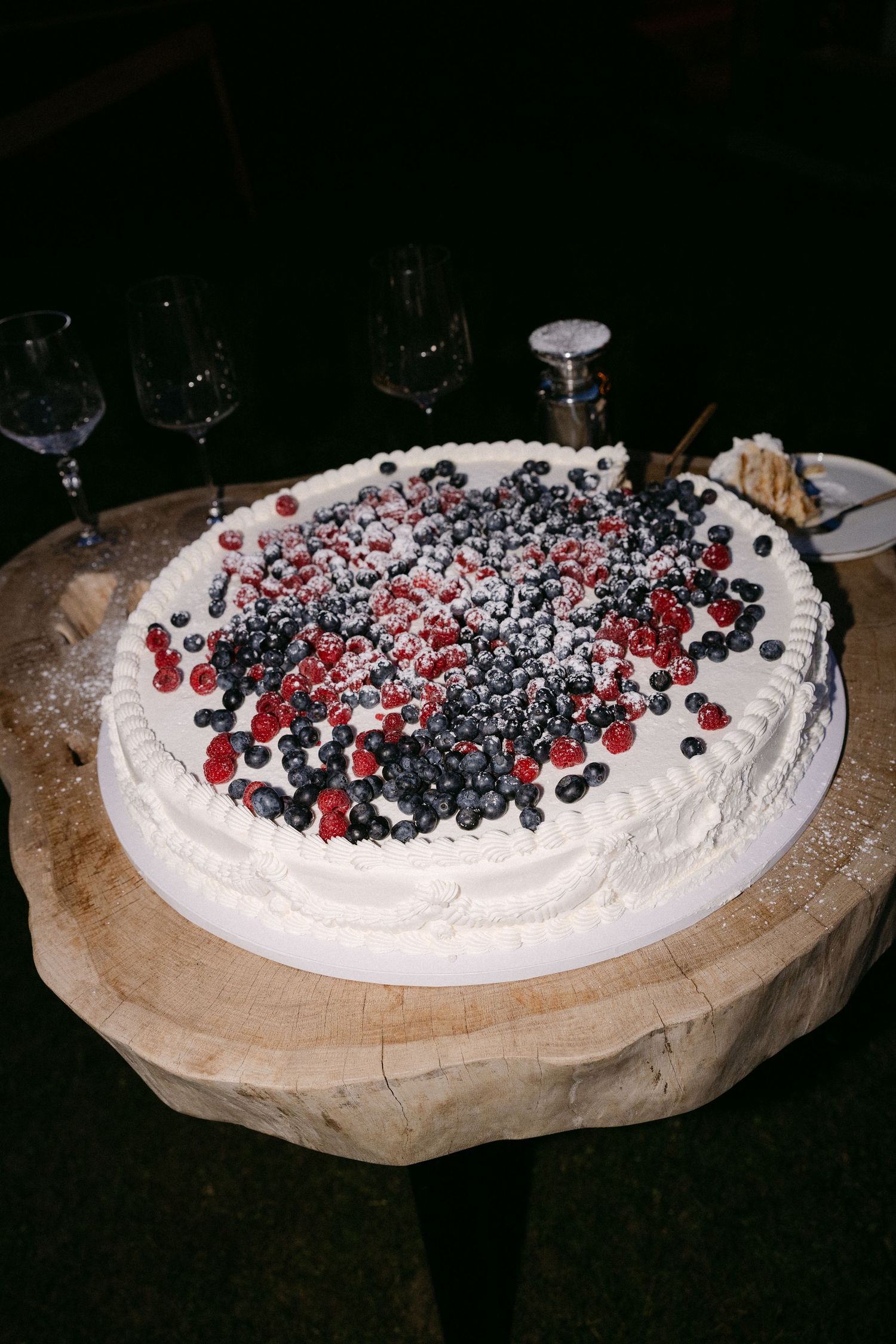 White wedding cake topped with fresh blueberries and raspberries dusted with powdered sugar on a wood stand.