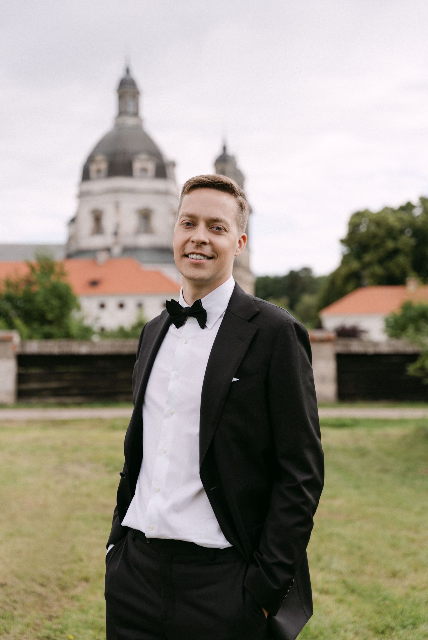 Smiling groom in black tuxedo with bow tie posing in front of a historic domed church building.