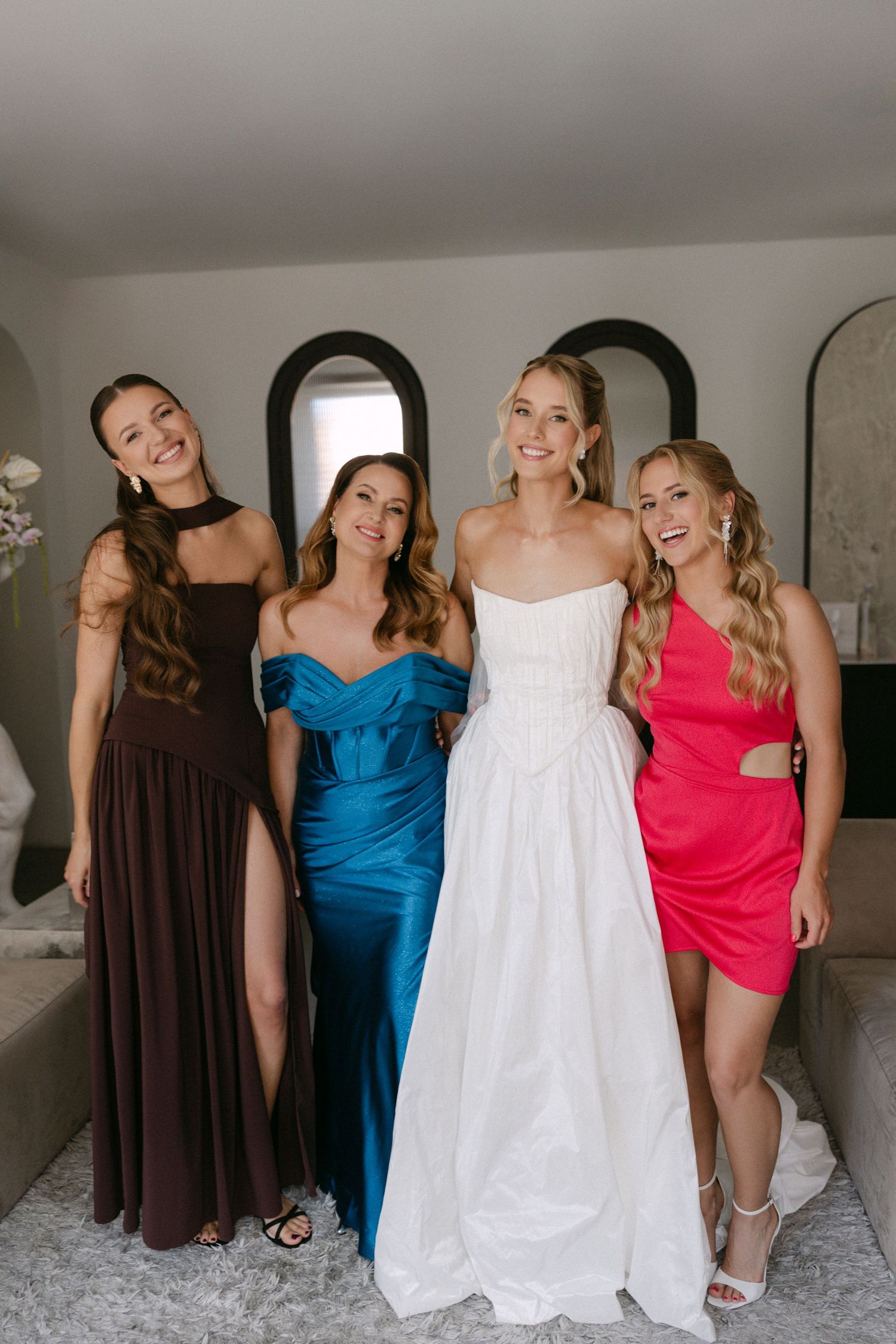 Bride in white gown poses with three women in colorful formal dresses before the wedding.