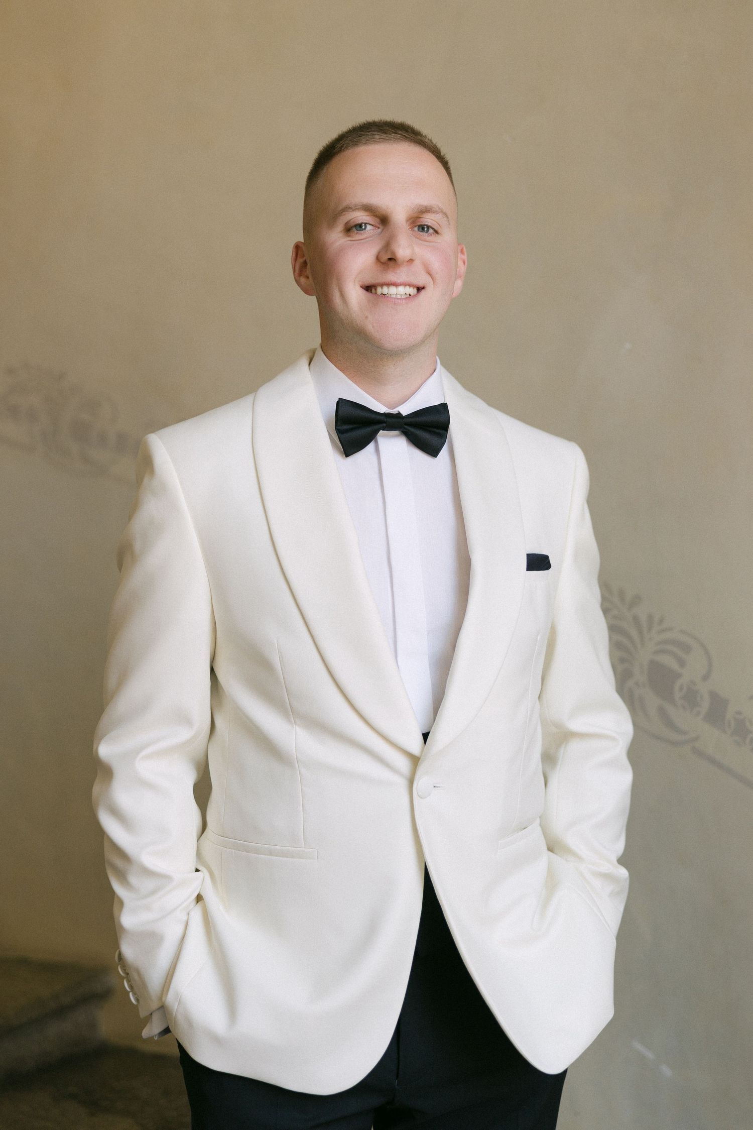 Smiling groom in white tuxedo jacket with black bow tie and pocket square posing indoors.