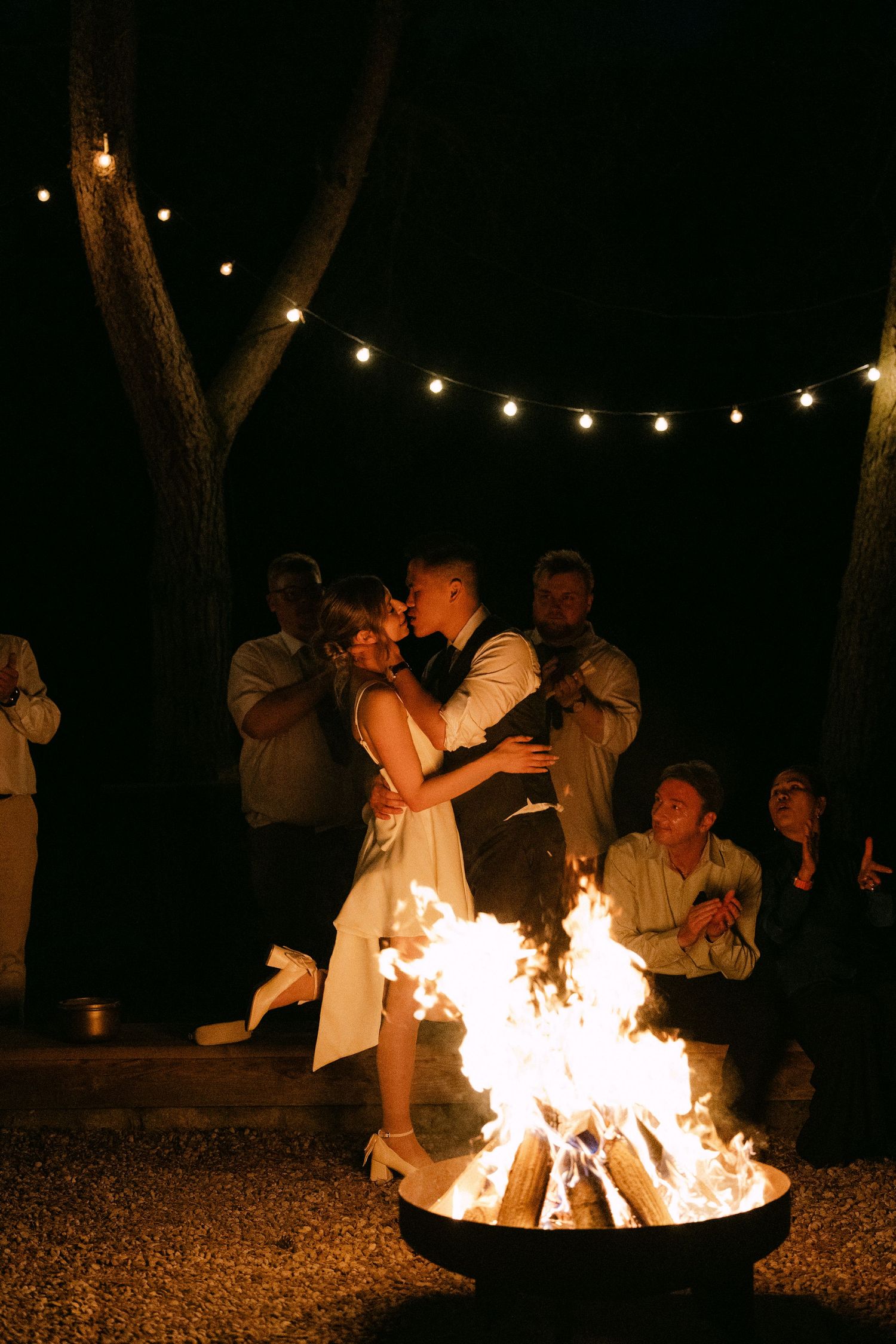 Newlyweds share a romantic kiss by a glowing fire pit under string lights surrounded by guests at night.