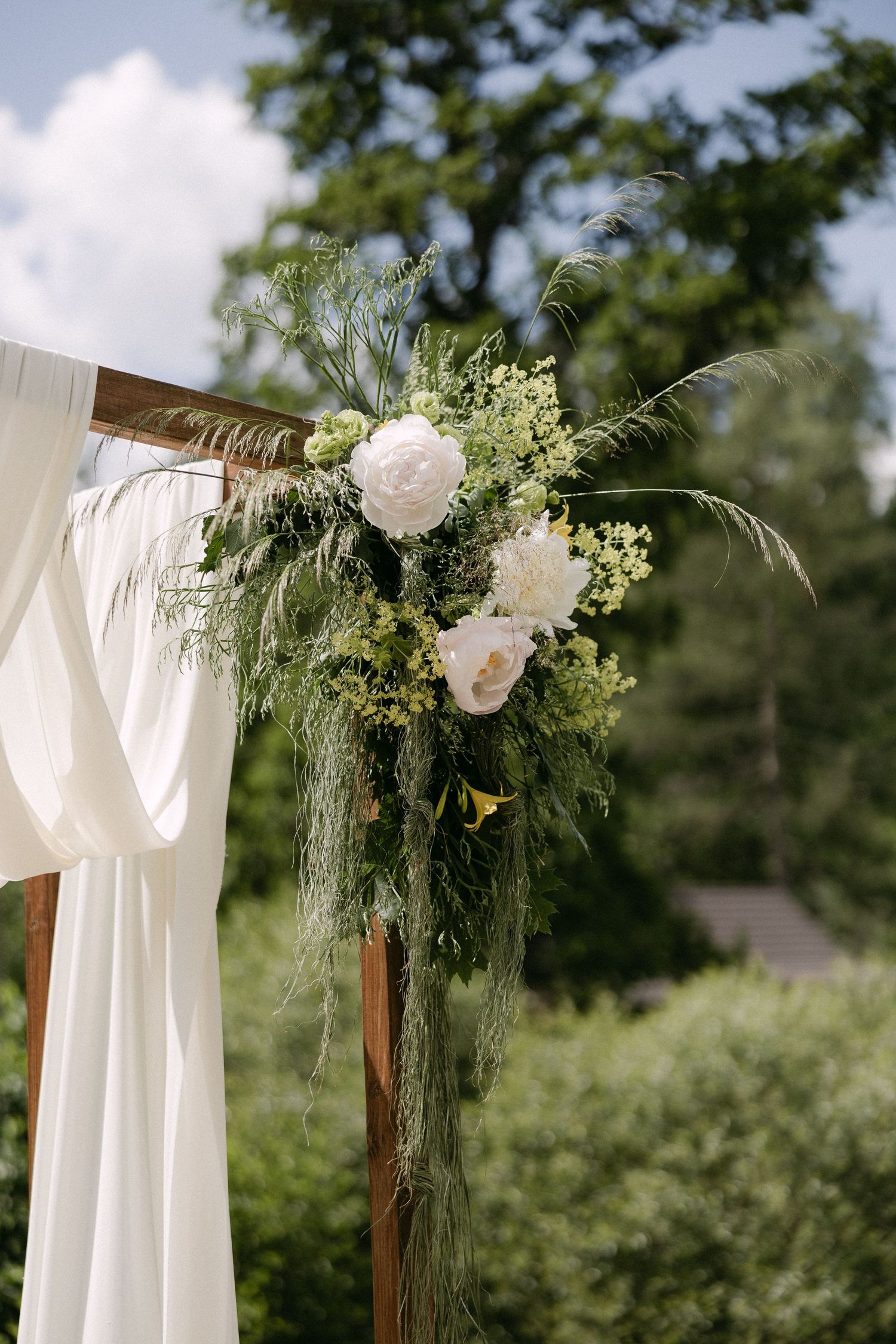Rustic wooden wedding arch adorned with white peonies and lush greenery with flowing white fabric outdoors.