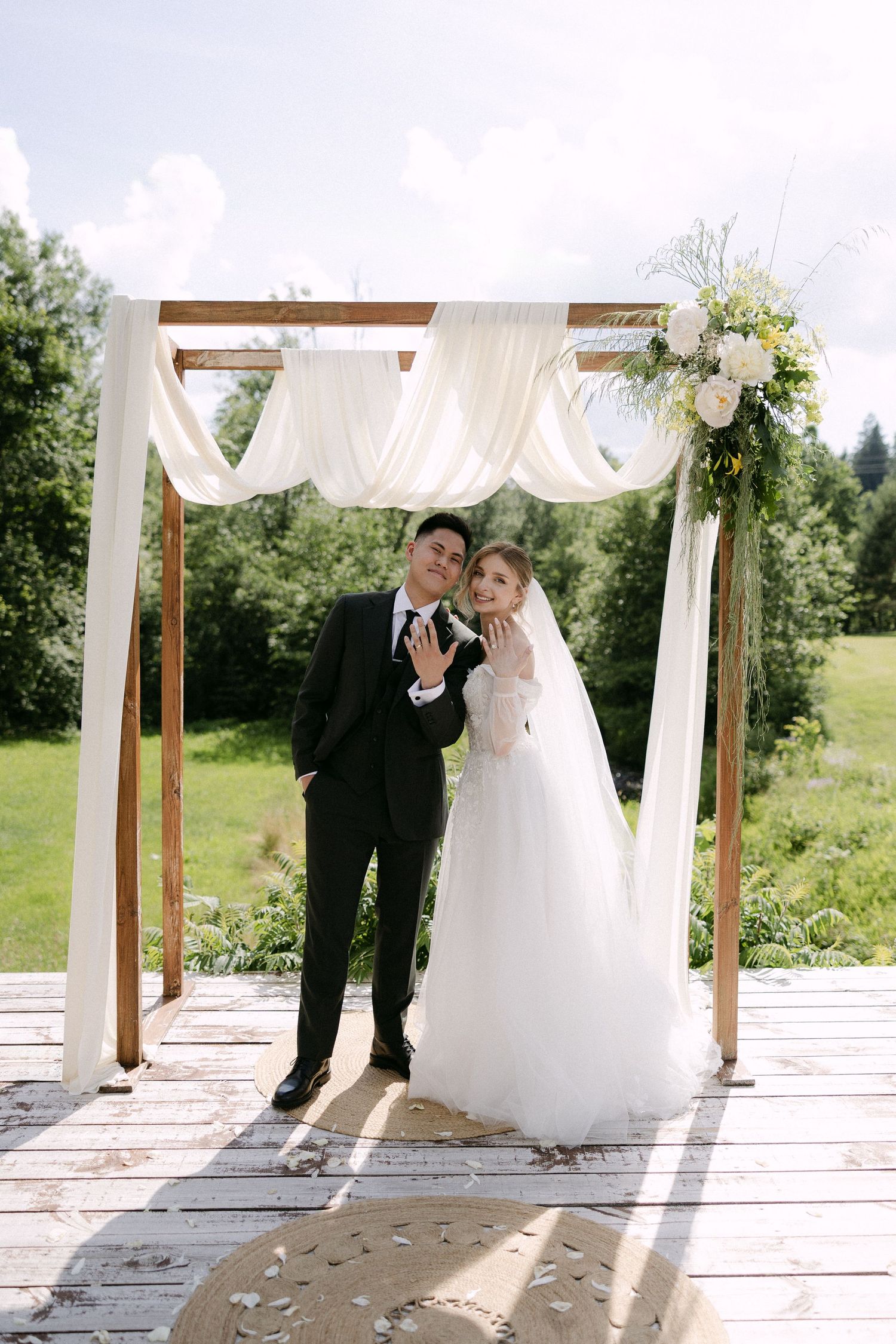 Newlyweds show wedding rings while posing under floral arch at outdoor ceremony.