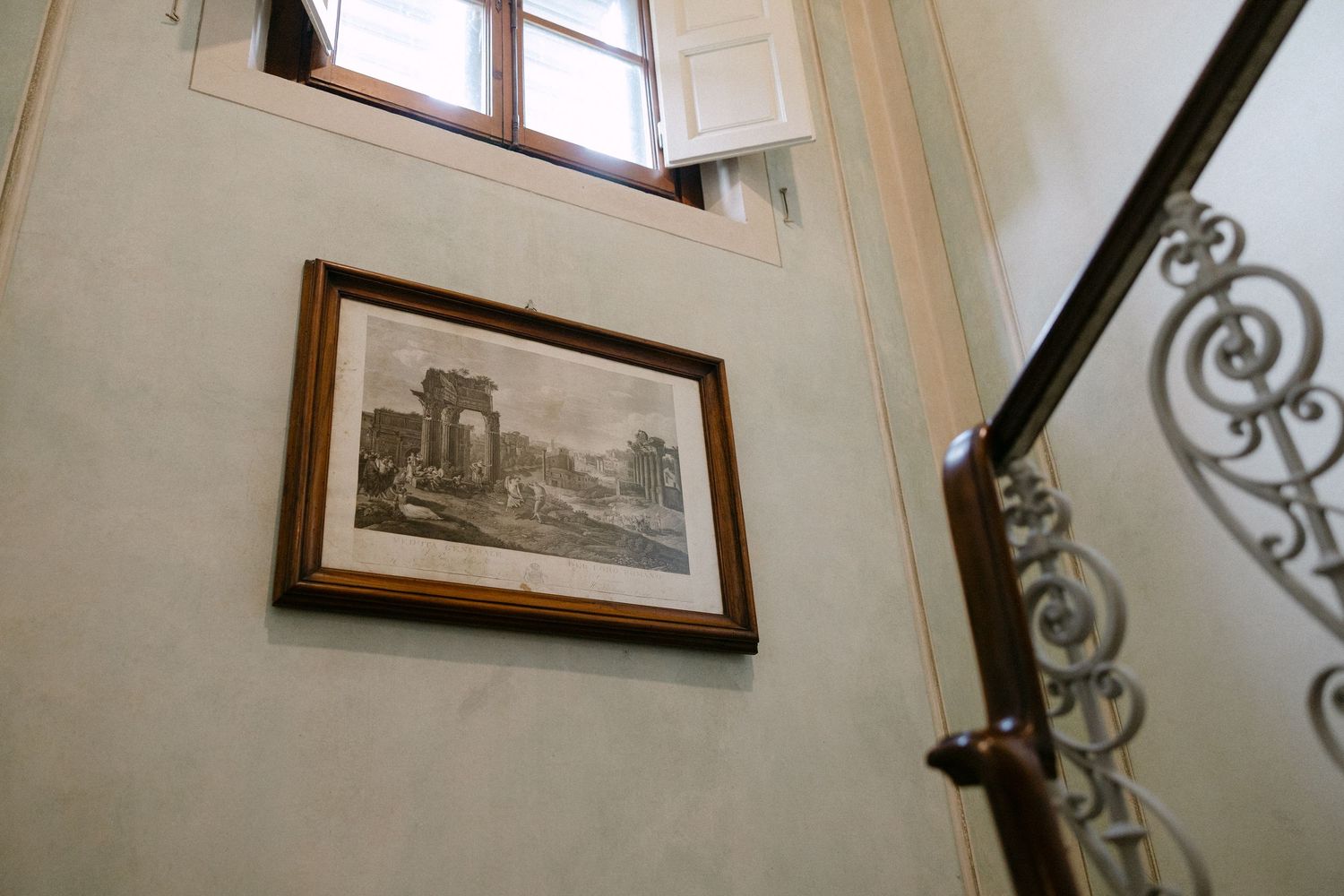 Framed black and white architectural print hanging on a pale green wall near an ornate wrought iron staircase railing.