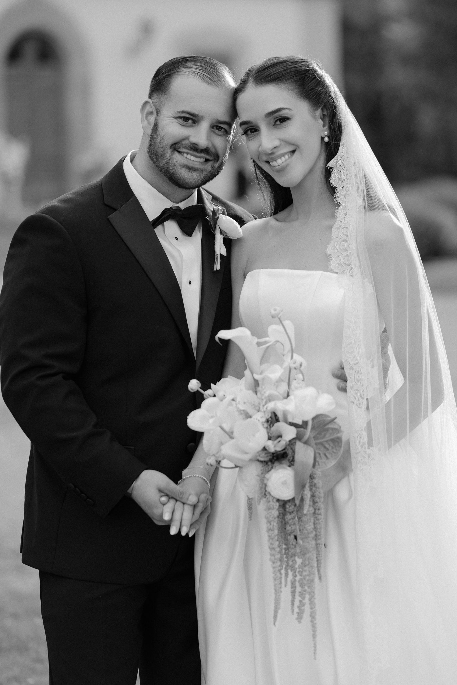 Black and white portrait of smiling bride and groom holding white floral bouquet on their wedding day.