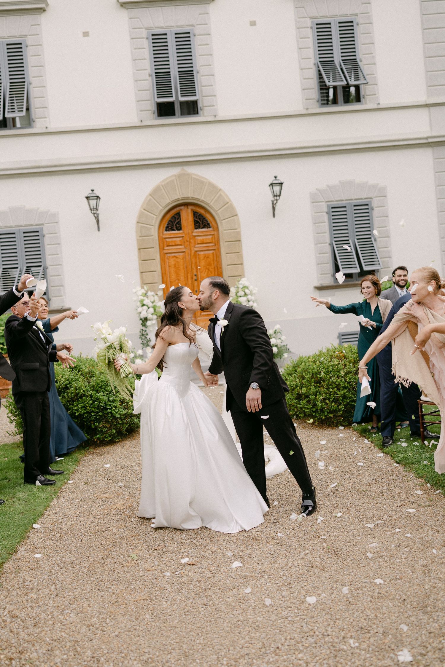 Newlyweds share a kiss as guests toss white petals outside a classic Italian villa wedding venue.