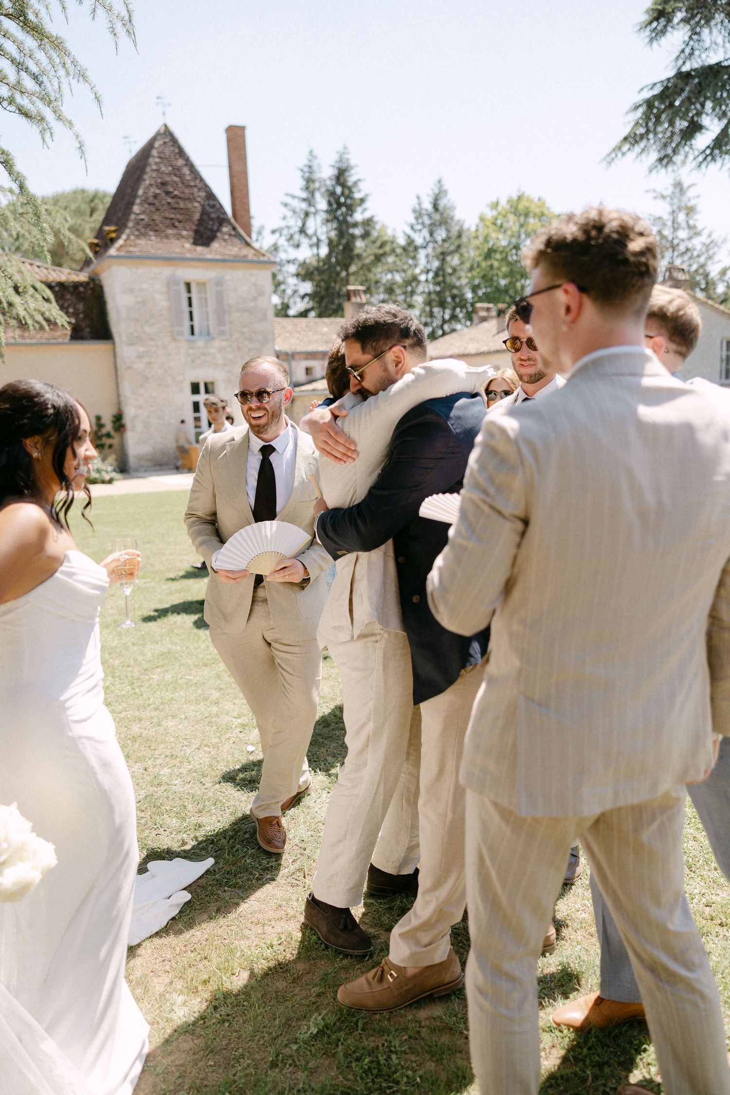 Groomsmen in beige suits hugging and celebrating at an outdoor wedding reception near a French chateau.