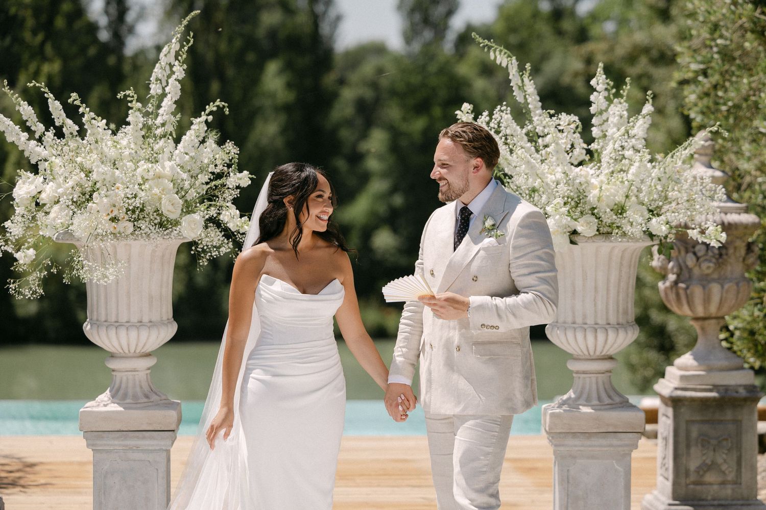 Bride and groom walk hand in hand past white floral urns at outdoor wedding ceremony by a pool.